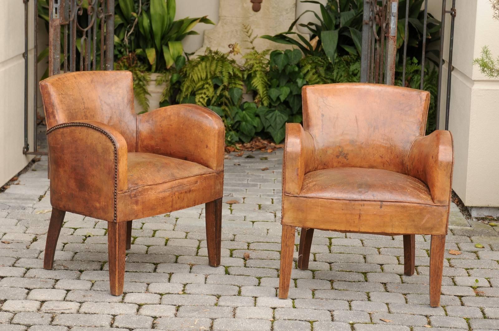 A pair of English leather club chairs with nailheads from the early 20th century. Each of this pair of English armchairs features a brown leather covered body with nailhead trim, following the outline of the rounded arms and the back. The slightly