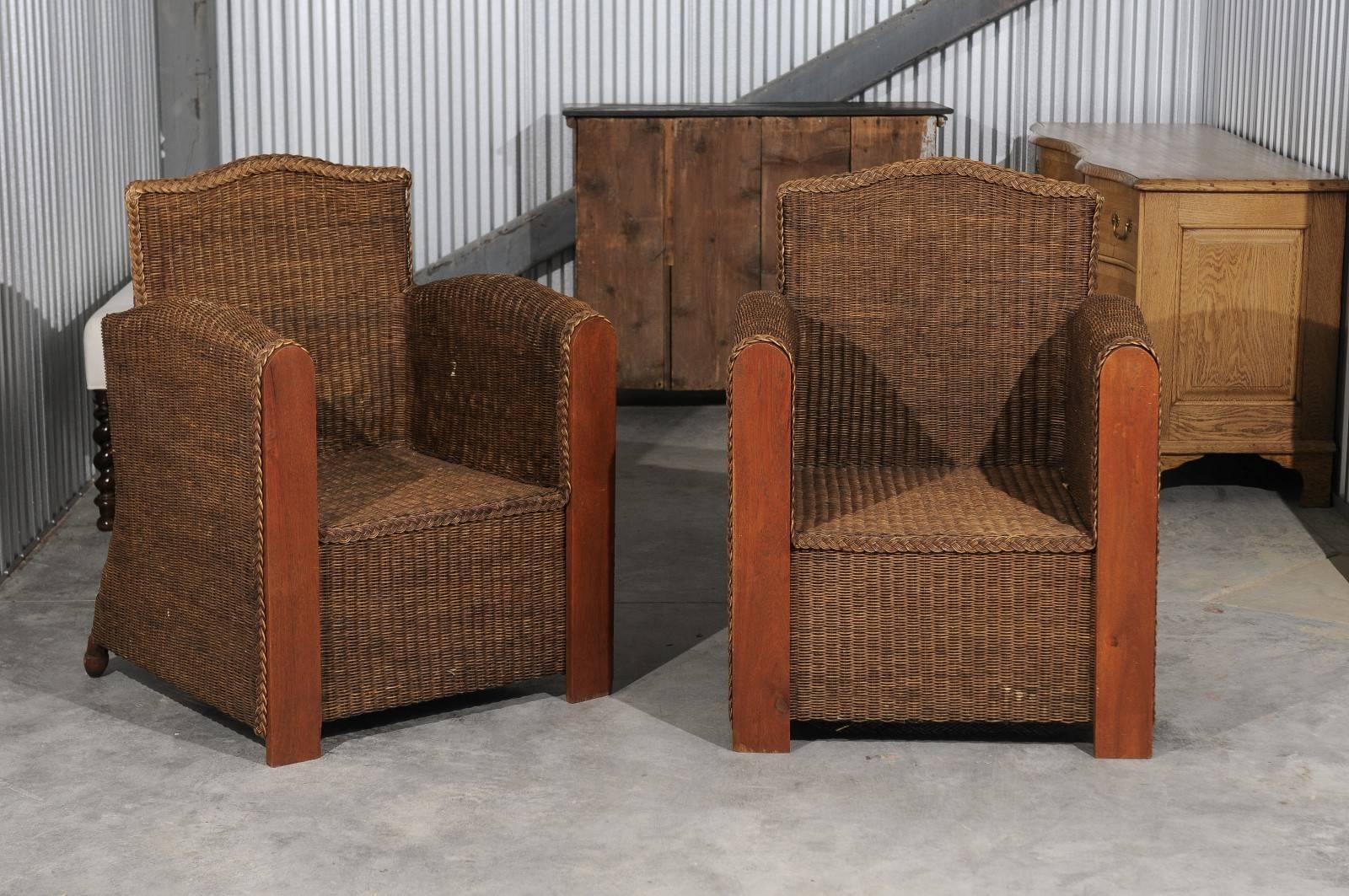 A pair of French vintage wicker and wood club chairs from the mid-20th century. Featuring a wicker body with wooden arm supports, each chair is adorned with a straight camel back. The tall straight arms provide a greater sense of privacy and the
