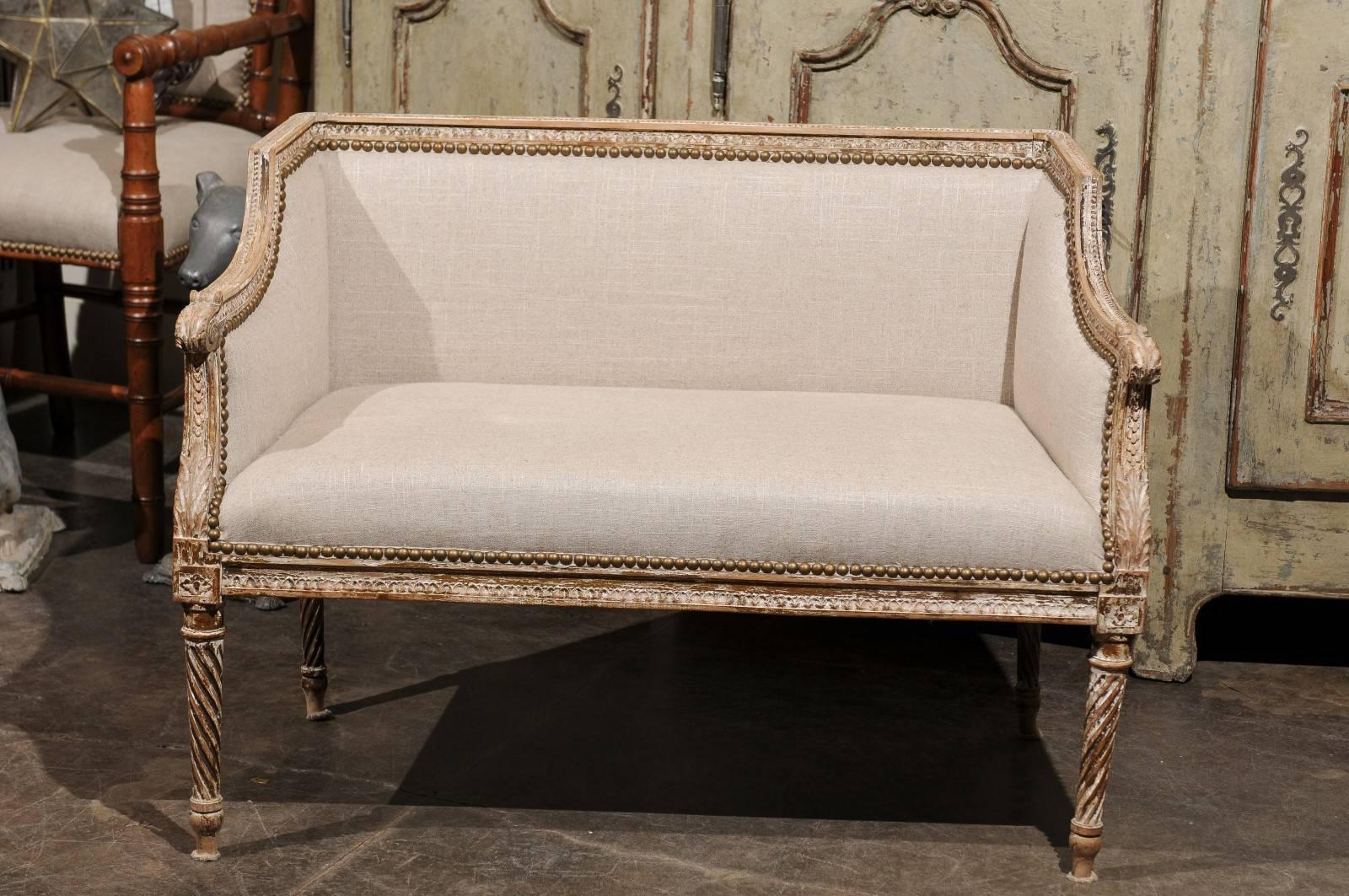 A French Louis XVI style petite wooden bench with distressed paint and linen upholstery from the 1820s-1840s. This French small bench features a straight back and scrolled arms framing a rectangular seat. The back, solid arms and seat are covered in