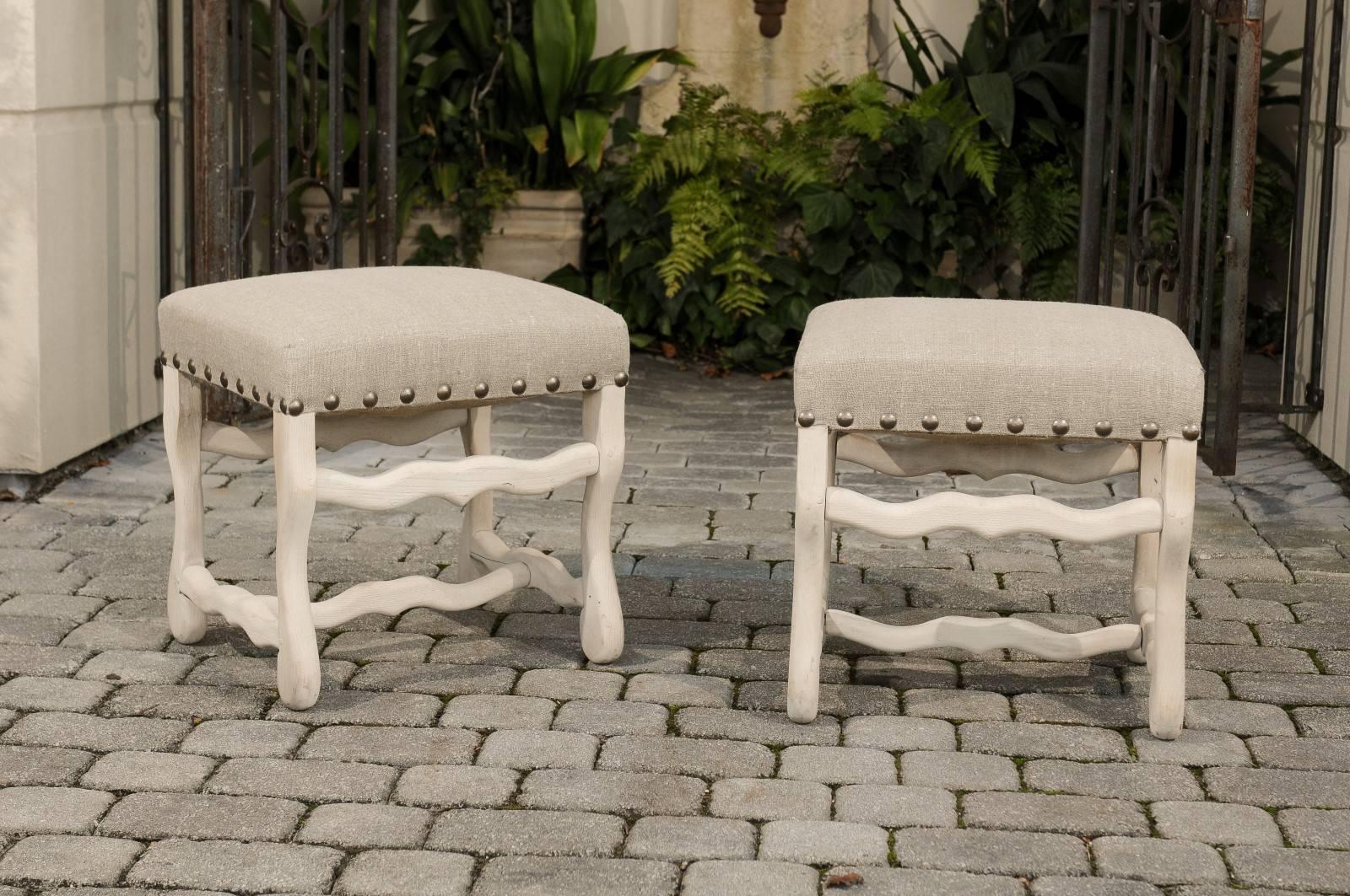 A pair of French Louis XIII style bleached wood upholstered os de mouton stools from the Turn of the Century. Each of this pair of carved stools features a rectangular top covered in linen upholstery, accented with a nailhead trim. Each stool sits