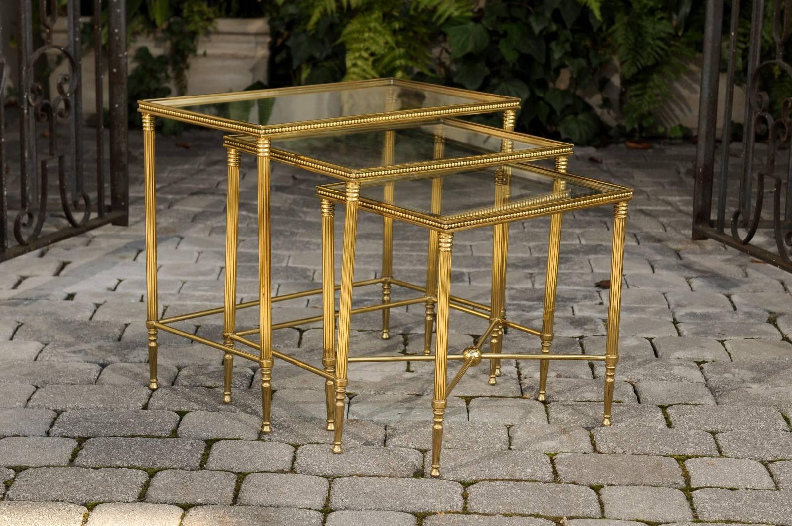 A set of three French nesting tables with glass tops and brass armature from the mid-20th century. Each of this charming set of French vintage nesting tables features a 1.75 thick glass top adorned in its surround with a mirrored frame and supported