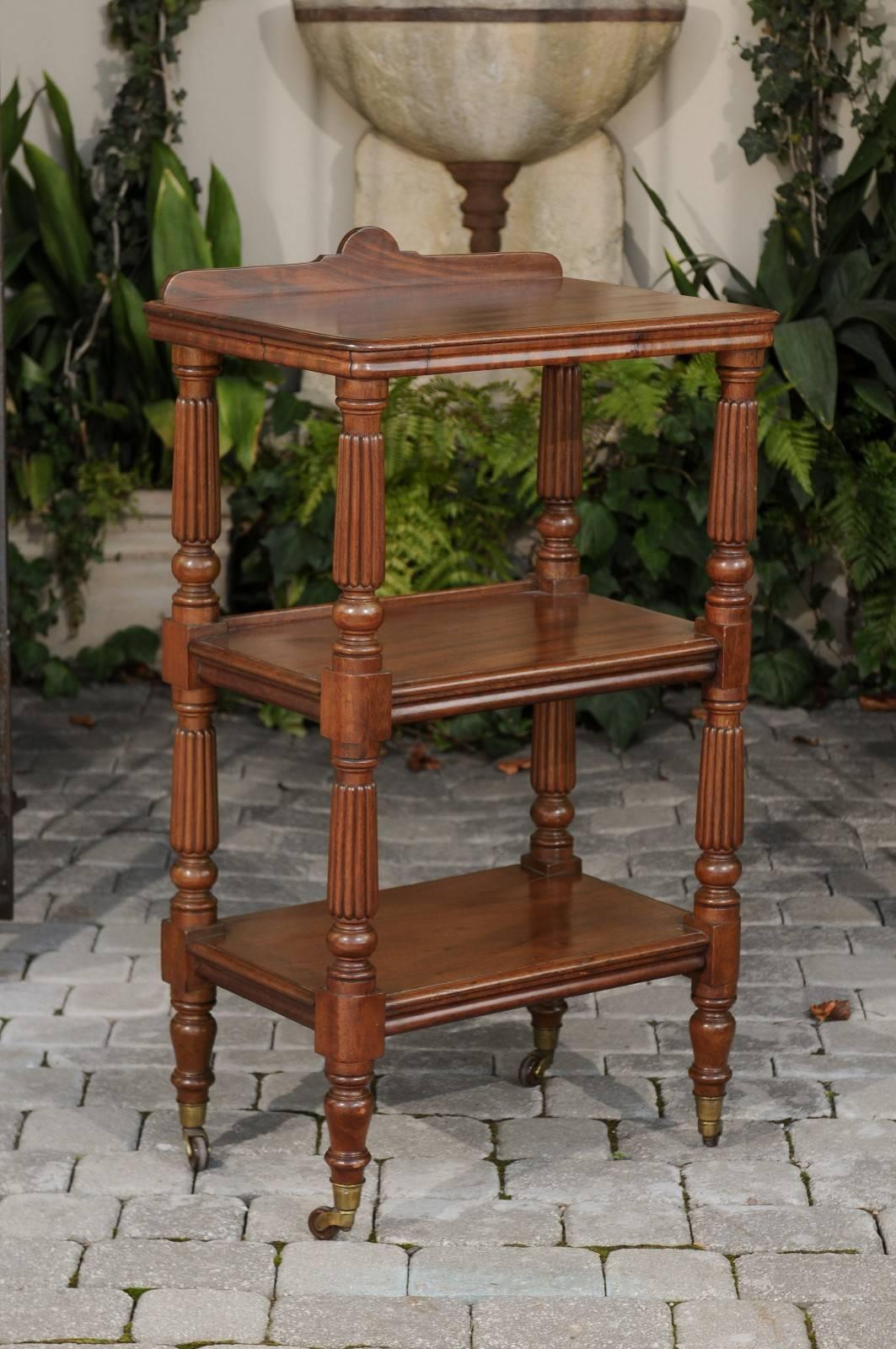 An English narrow three-tiered walnut trolley on casters from the second half of the 19th century. This English wooden trolley features a rectangular top with moulded edges and raised back, resting on four turned and fluted posts. These posts