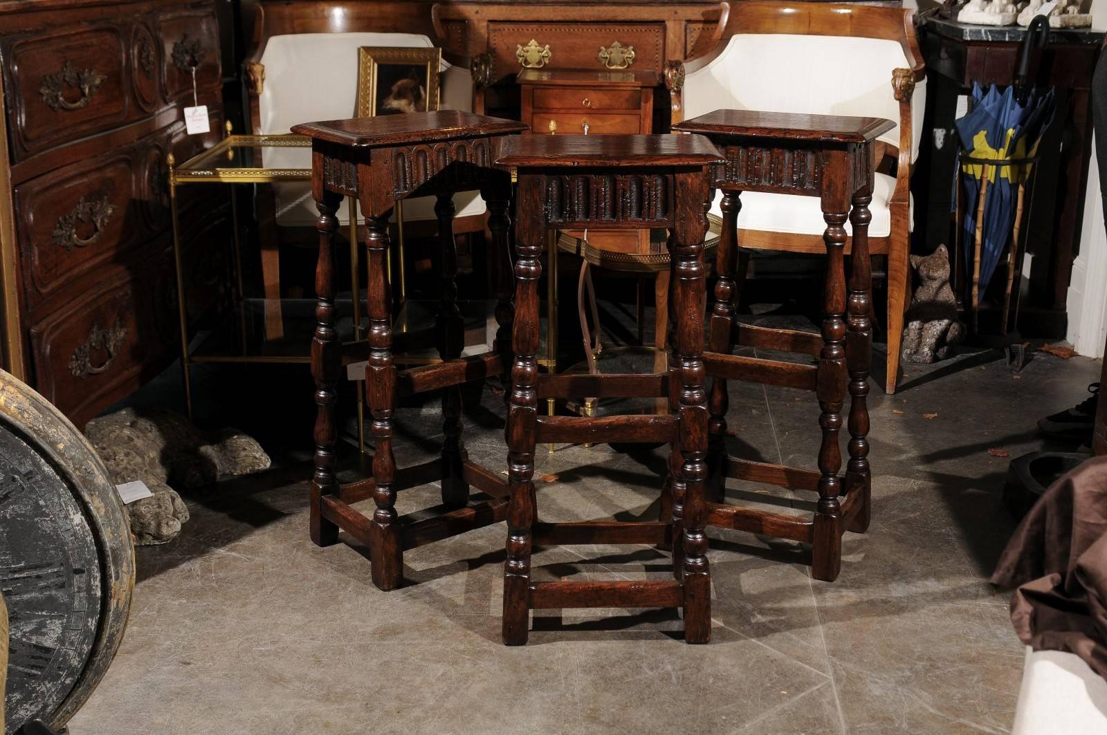 A set of three English Ipswich oak bar stools from the early 20th century. Each of this set of three wooden stools features a rectangular top with bevelled edges sitting above a nicely carved apron adorned with scoop patterns on all sides. The seats