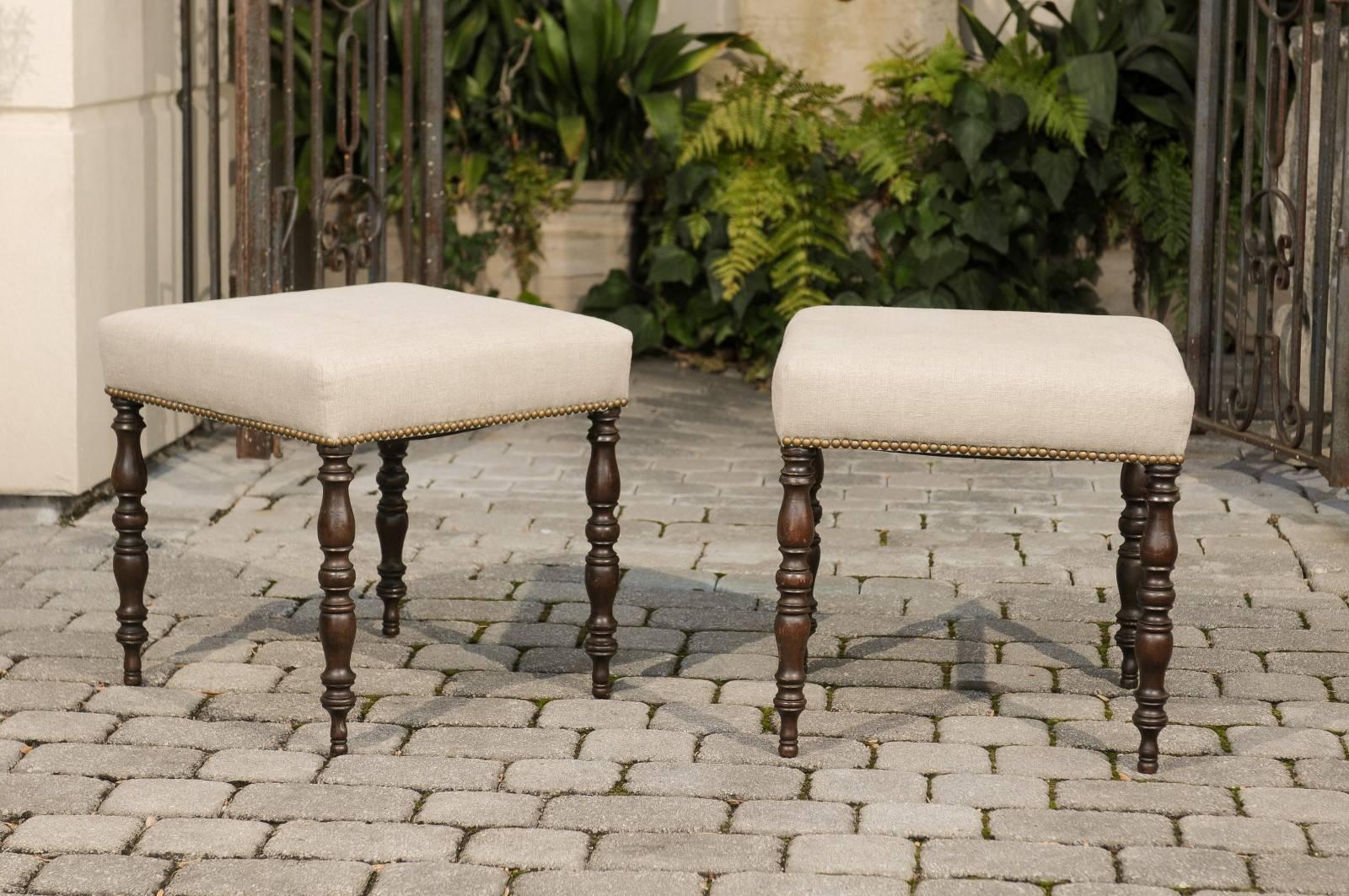 A pair of English, late 19th century walnut stools with turned legs and upholstered seats with brass nailhead trim. Each of this pair of walnut stools features a seat newly recovered in a simple linen fabric with nailhead trim. The ensemble is