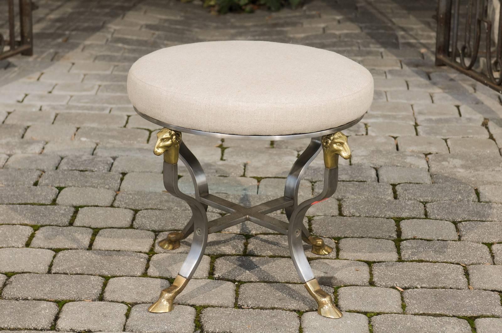 An Italian Directoire style steel and brass upholstered stool with ram's heads and hoofed feet from the mid 20th century. Born in the 1950s, this Italian stool features a round seat that has been recovered with a linen fabric. This simple looking