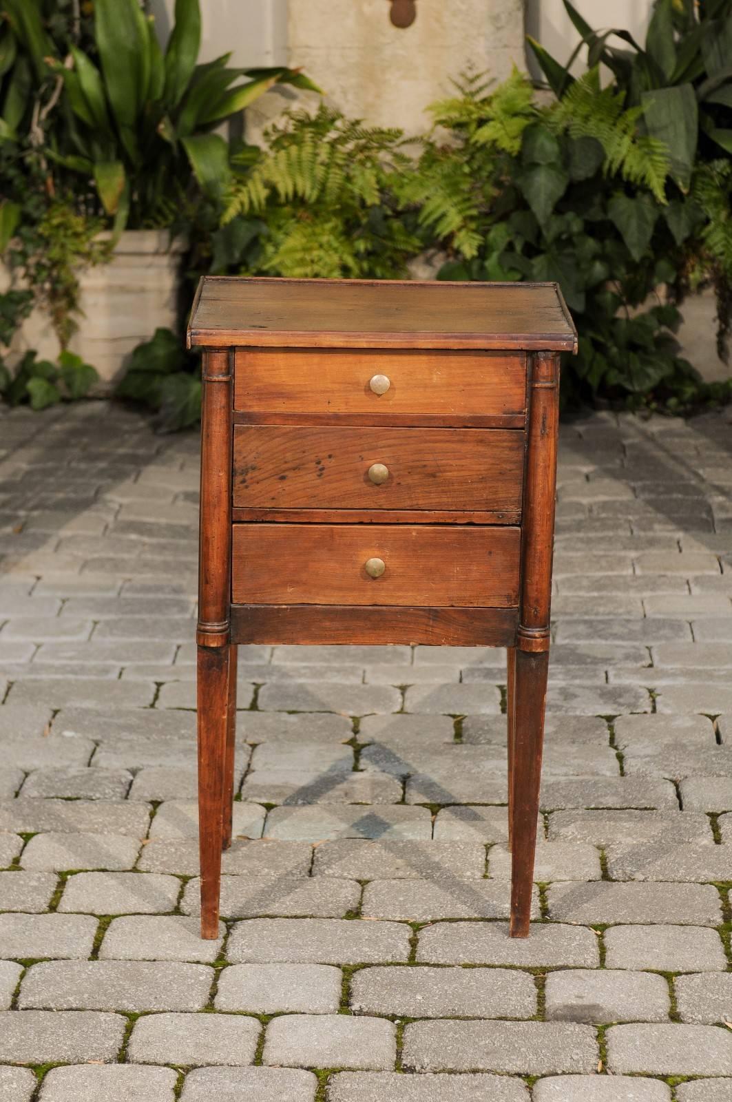 A French petite walnut Neoclassical commode with three drawers, semi-columns and tapered legs from the early years of the 19th century. This French petite commode features a rectangular top surrounded by a flat molding on three sides, sitting above