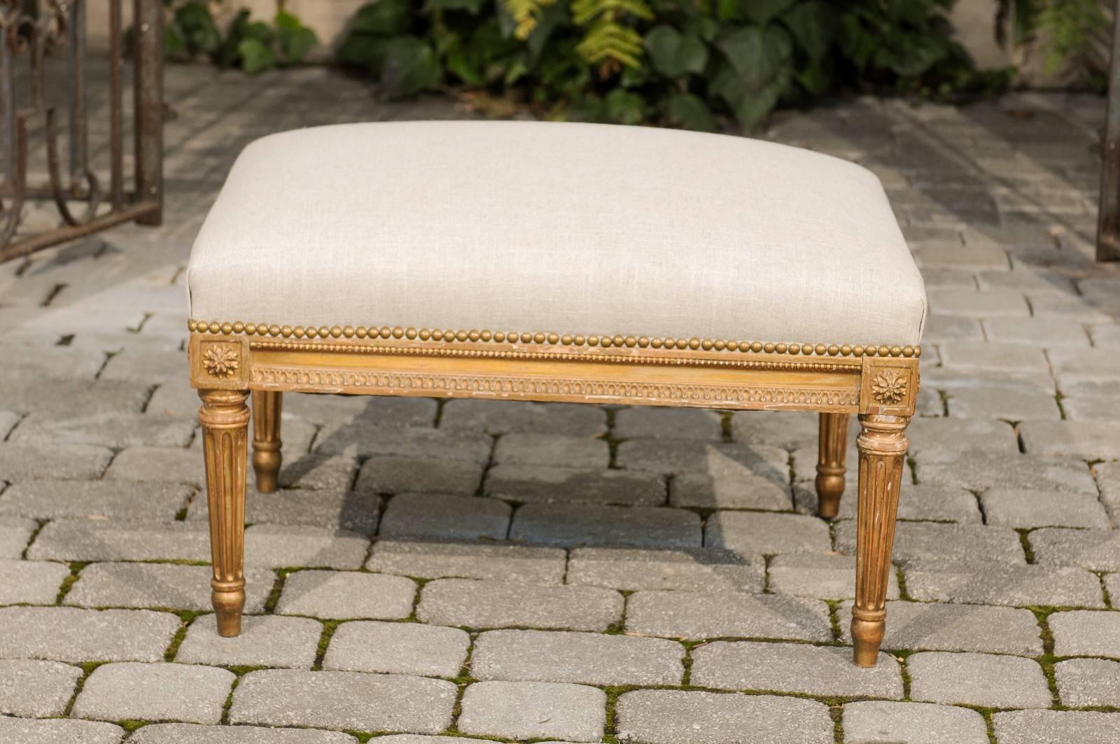 A French Louis XVI style giltwood ottoman from the early 20th century with upholstered seat and nailhead trim. This French Louis XVI style ottoman features a rectangular seat newly recovered in a simple linen fabric with brass nailhead trim. The