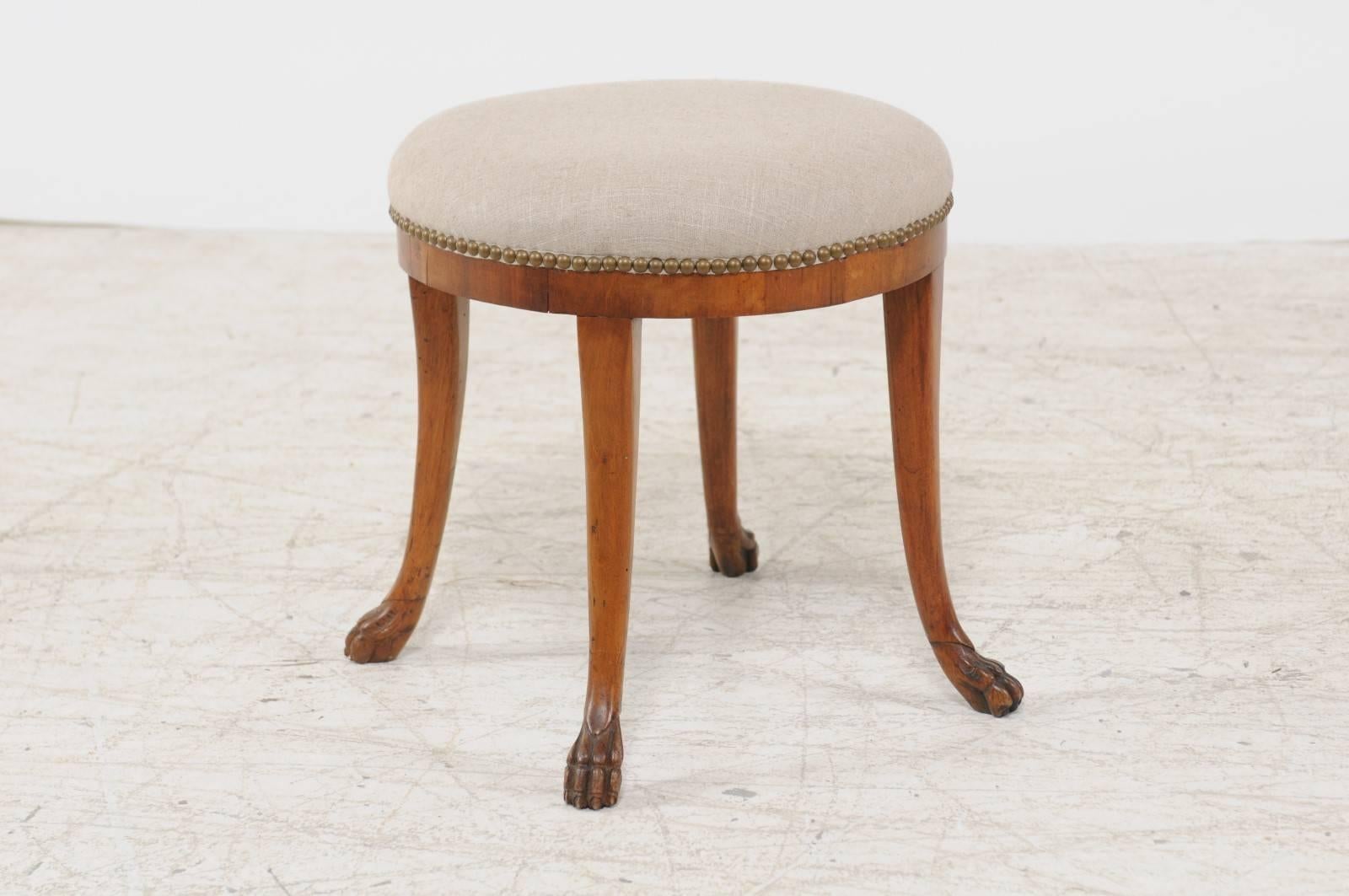 An Austrian Biedermeier round stool with saber legs and lion paw feet from the mid 19th century and new linen upholstery. This Austrian stool features a circular top, recovered with a new linen upholstery. The stool is raised on four saber legs
