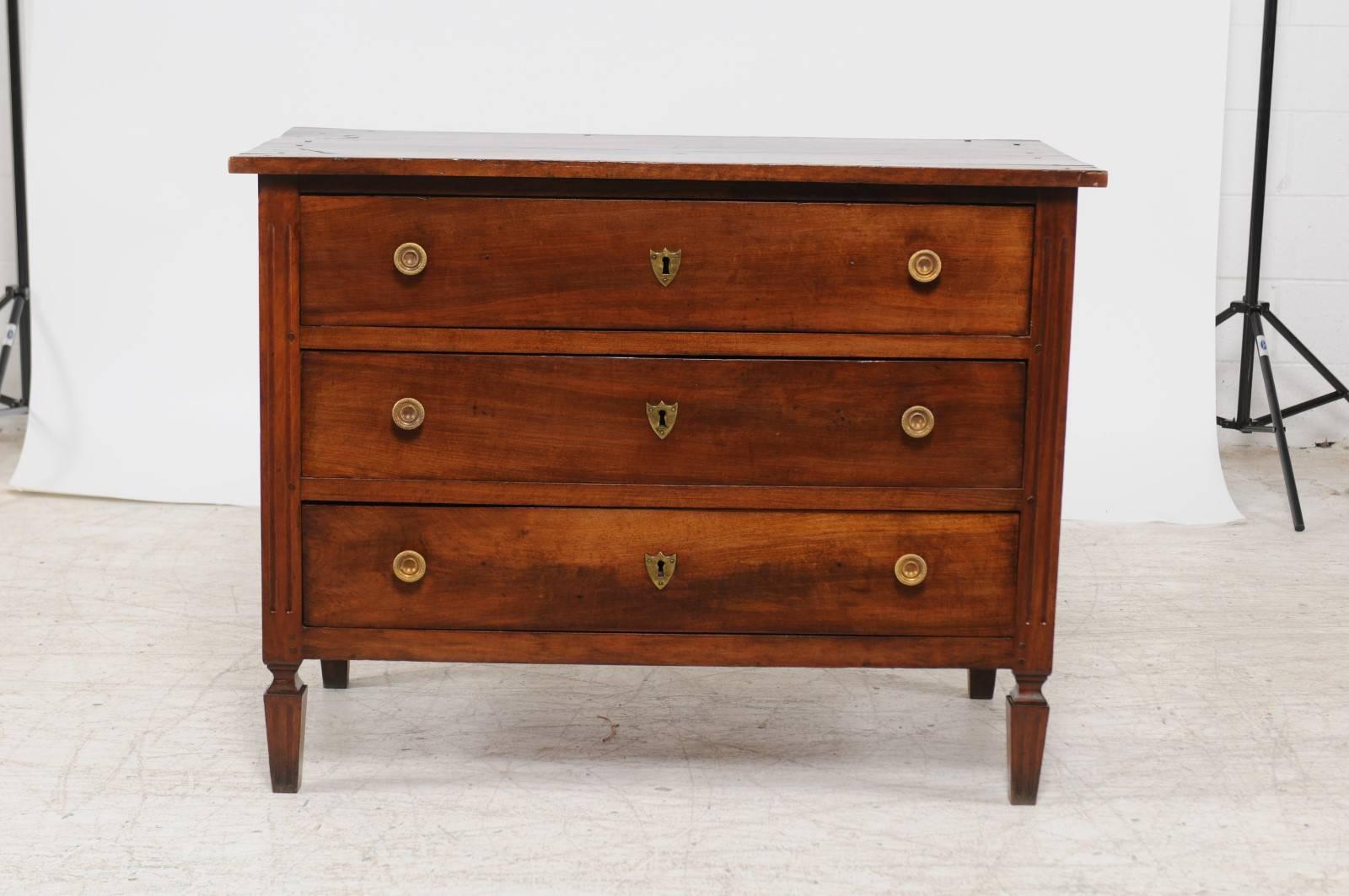 An Italian neoclassical walnut three-drawer commode with tapered legs from the early 19th century. This Italian wooden chest-of-drawers features a rectangular planked top sitting above three dovetailed drawers. Each drawer is simply adorned with