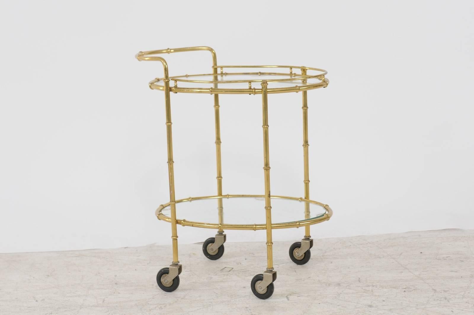 A French vintage brass cart from the 1950s with glass oval shelves and casters. Born during the mid-20th century, this exquisite brass cart features two levels of oval glass shelves. The upper section of the piece showcases a curvilinear handle