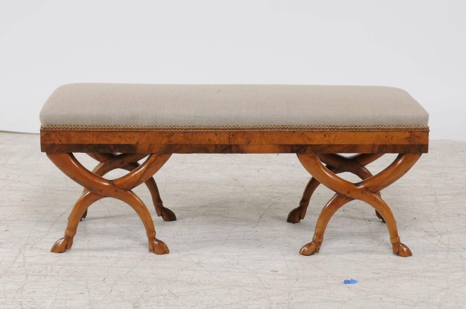 An Austrian Biedermeier double X-frame bench with burl veneer and hoofed feet from the mid-19th century and new upholstery. This exquisite Biedermeier bench features a rectangular linen upholstered seat with brass nailhead trim, sitting above a burl