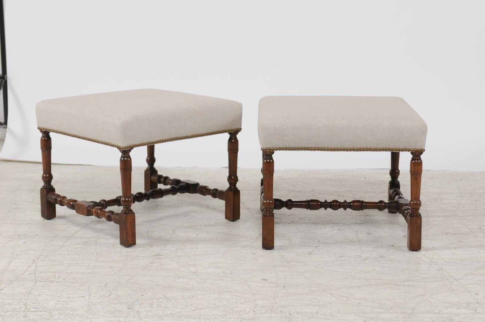 A pair of English walnut stools or ottomans with turned legs and cross stretchers from the second half of the 19th century and new upholstery. Each of this pair of English stools features a square seat, recovered with a simple linen upholstery