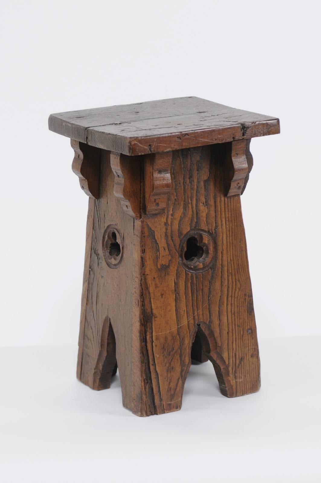 A rustic Spanish oak square-shaped stool with trefoil motifs from the early 20th century. Born in the early 1900s in Spain, this small wooden stool features a square planked top supported by elegant brackets and sitting above a solid base adorned