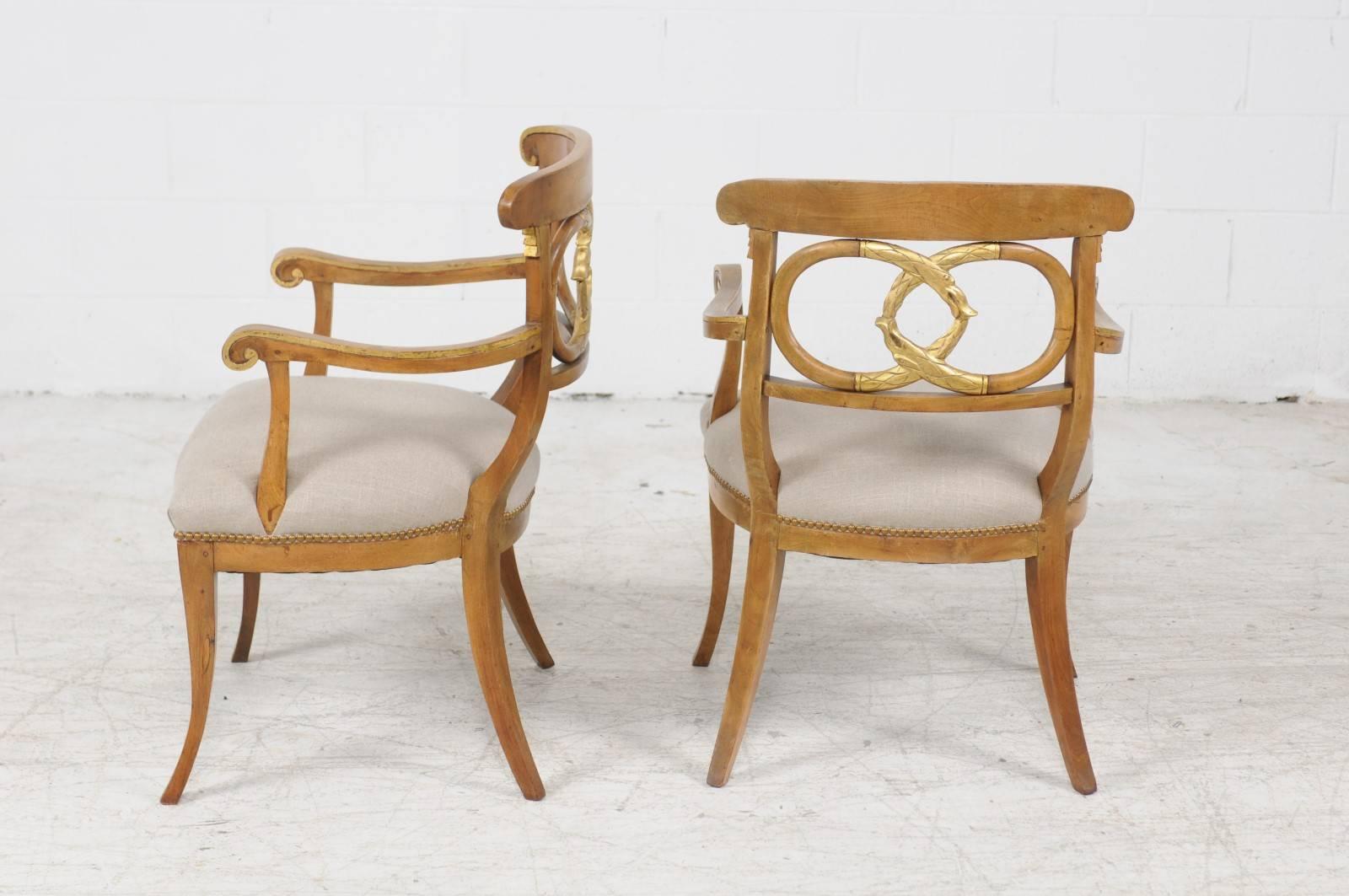 A pair of Italian parcel-gilt walnut armchairs with intertwined serpents and saber legs from the mid 19th century and new upholstery. Each of this pair of Italian chairs features a curved back with gilded accent on the upper rail. The eye is
