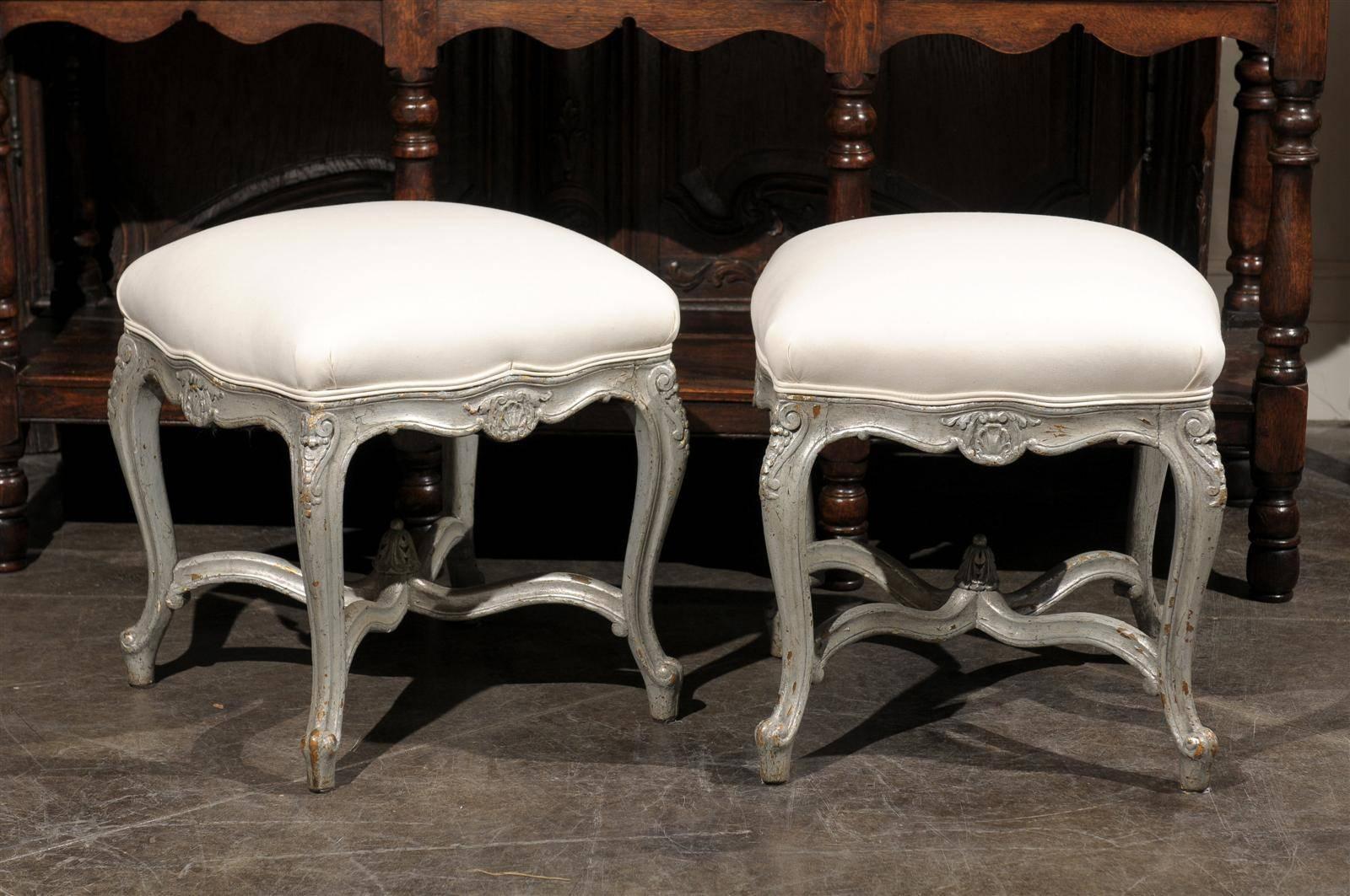 This pair of French Louis XV style silver leaf upholstered stools from the early 20th century features muslin covered seats over nicely scalloped skirts with central carved motifs. This exquisite pair of French Louis XV style stools, dating from the