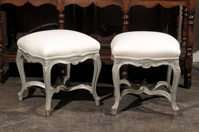 This pair of French Louis XV style silver leaf upholstered stools from the early 20th century features muslin covered seats over nicely scalloped skirts with central carved motifs. Each stool is raised on four cabriole legs with lovely volutes on