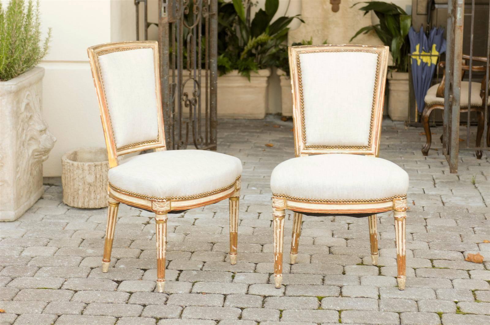 This exquisite pair of French Louis XVI style painted side chairs features slightly slanted upholstered backs over muslin upholstered seats with nailhead trim. The carved rosettes on the knees are rounded to follow the line of the tapered legs