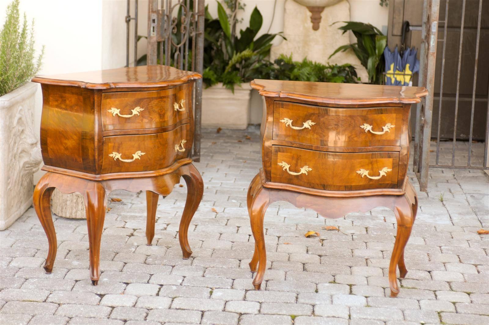 This pair of Italian Louis XV style walnut petite bombé commodes features for each piece a molded, shaped top over two serpentine drawers with slightly bowed sides. Two baroque style brass pulls adorn each drawer, nicely standing out against the