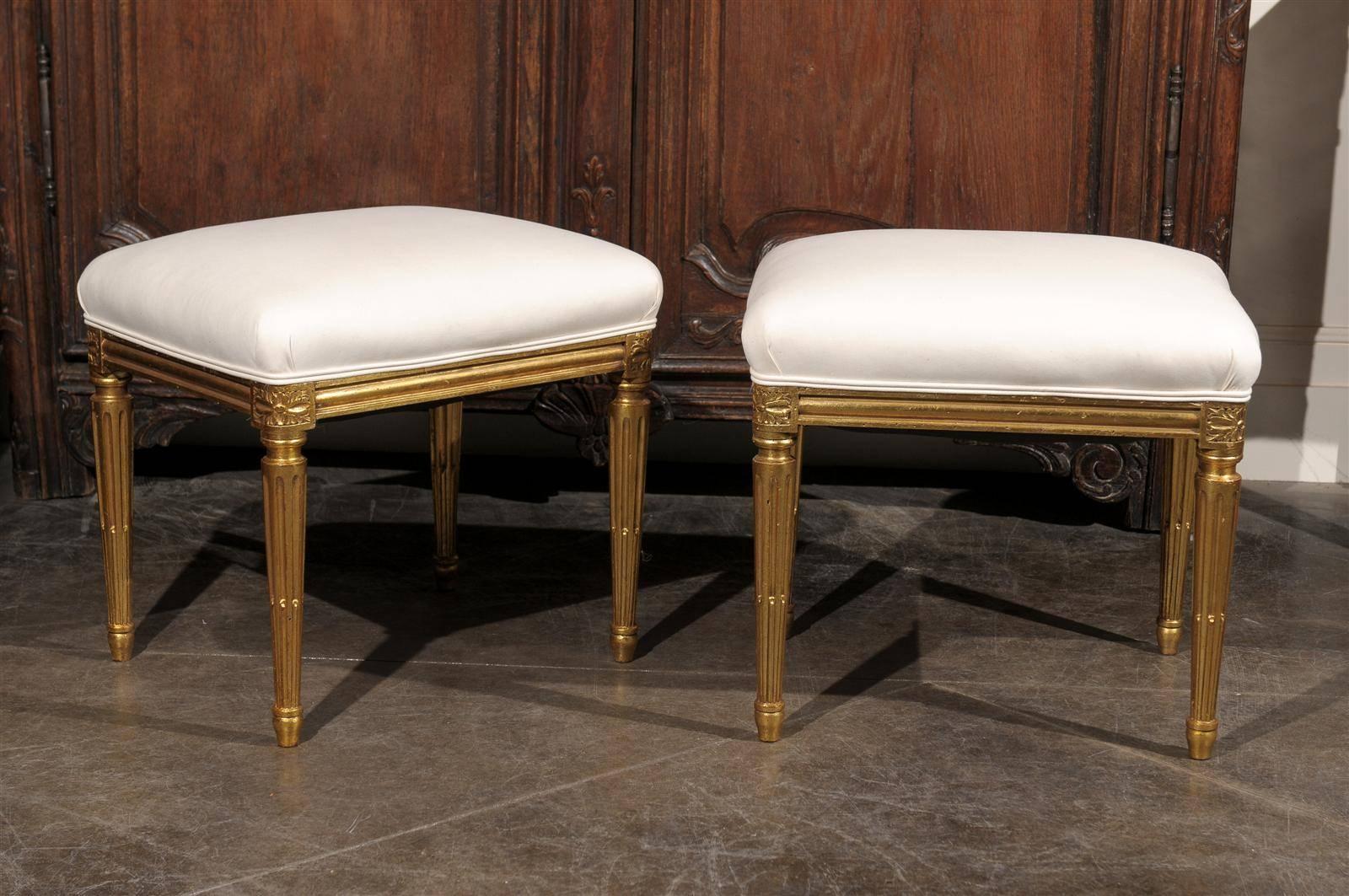 A pair of French upholstered and gilded stools from the early 20th century. This elegant pair of French Louis XVI style stools from circa 1920 features a new muslin upholstery applied on the rectangular seat. The body of the stools is made of gilded