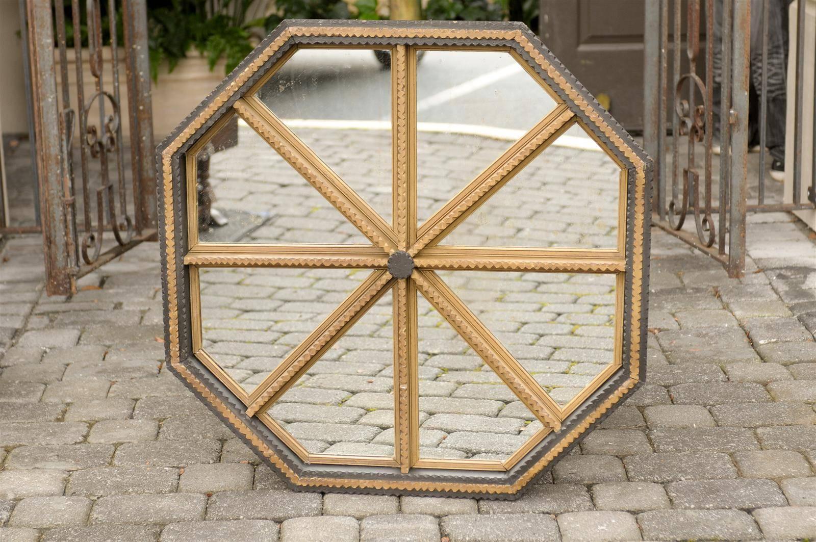 This American Tramp Art mirror from the mid 20th century features an octagonal frame painted in gold and black. The mirrored part is separated into sections, radiating from a black painted medallion in the center. The simple décor is made of carved
