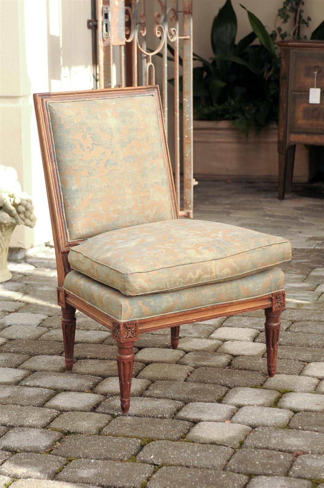 This French Louis XVI style upholstered slipper chair from the turn of the century (19th to 20th) features a rectangular slanted back over four fluted and tapered legs, adorned with small rosette motifs on the knees, typical of the Louis XVI style.