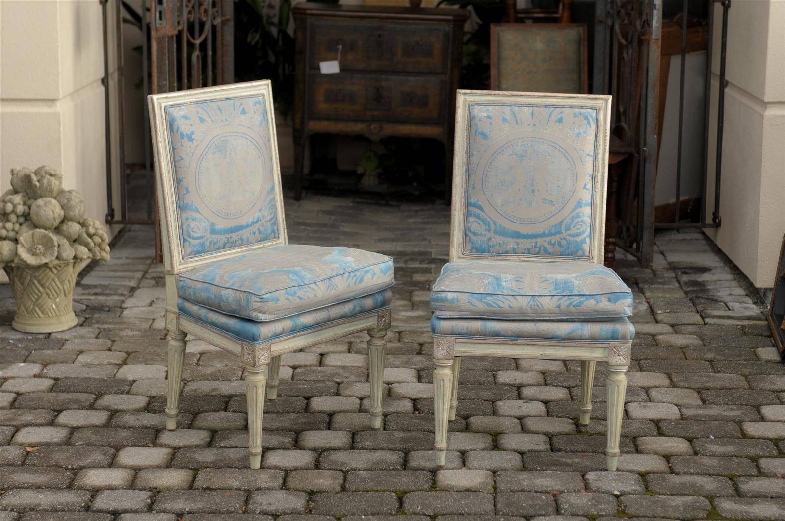 This pair of French Louis XVI style slipper chairs from the early 20th century features slightly slanted backs and seats upholstered with a blue themed Fortuny fabric. The light grey / green painted frames are highlighted with silver leaf accents.