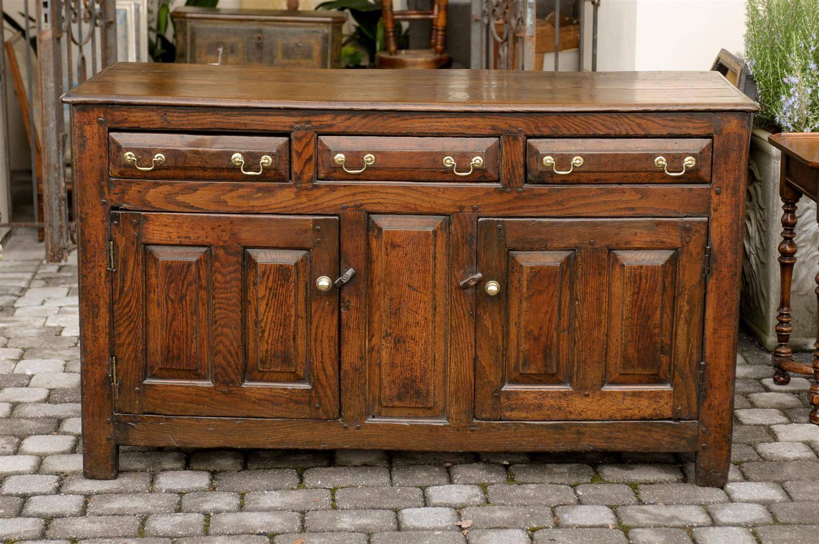 This English oak buffet from the very early part of the 19th century features three drawers over two doors. Each drawer is decorated with two brass bail handles. The two doors are symmetrically placed on each side of a raised panel, repeating this