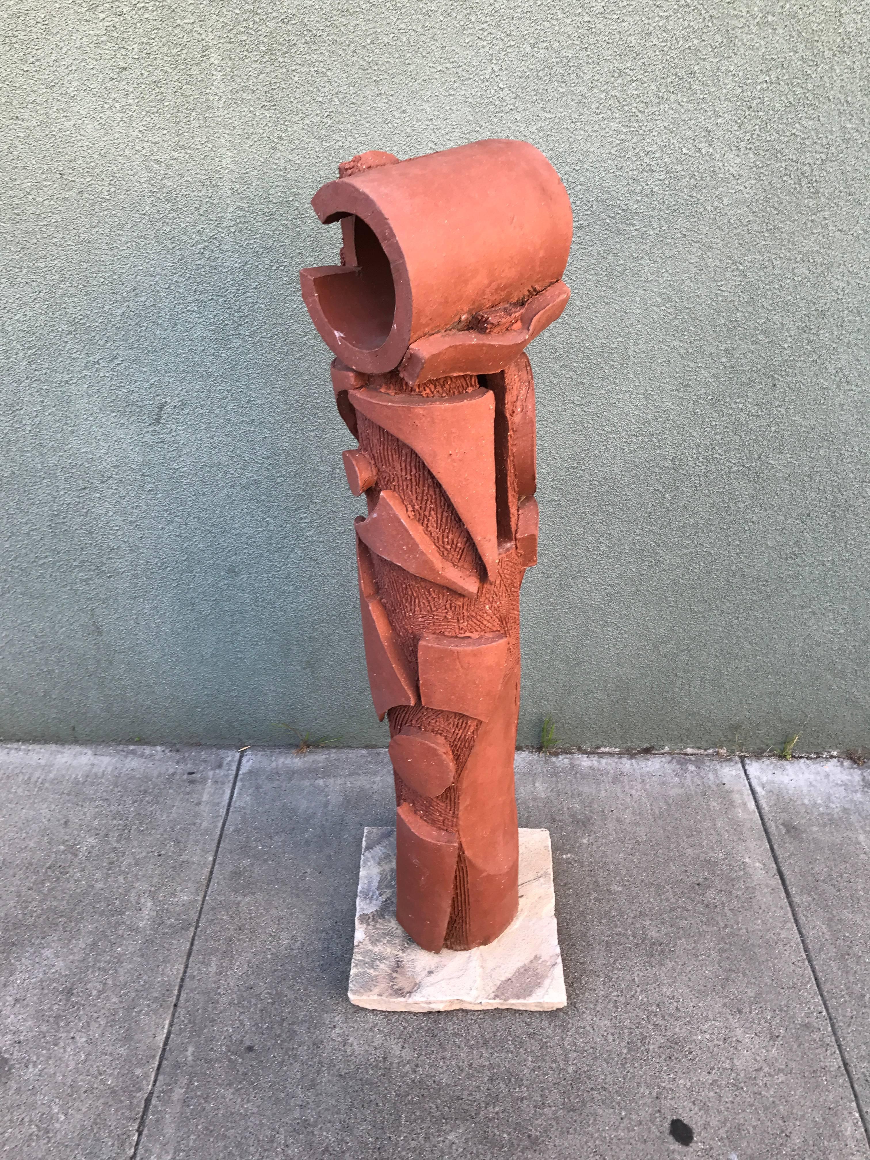 Large ceramic abstract or Brutalist sculpture in a natural red tone fired clay, a cylindrical form in which the clay has been carved to create both repeating form and texture on its surface. The base is a fossilized slate with ancient ferns embedded