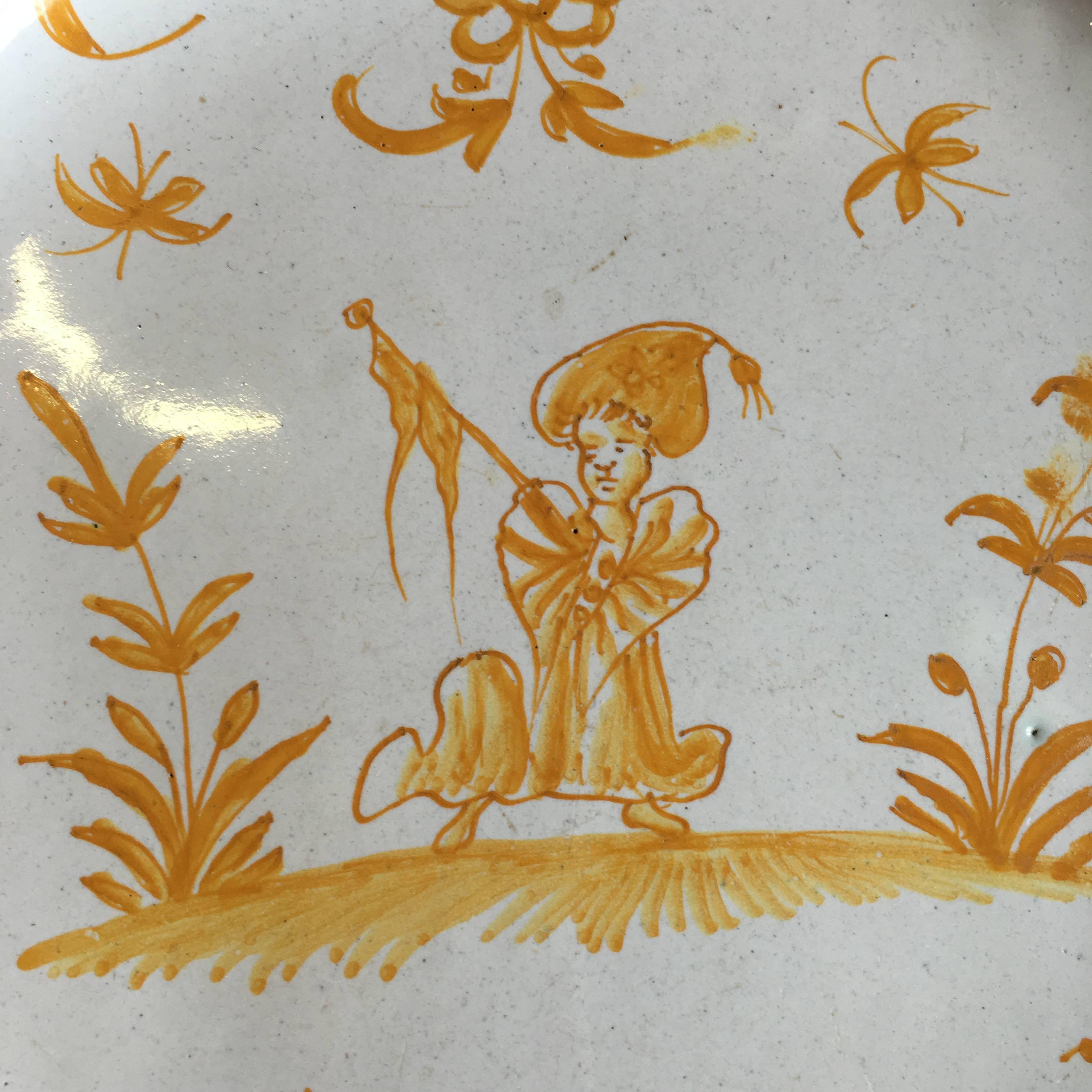 A charming 18th century French yellow glazed plate from Moustiers, circa 1780, depicting a stylized figure with a turban in a field with a flag. The plate has a scalloped edge and retains an early 19th century wall hanging devise.