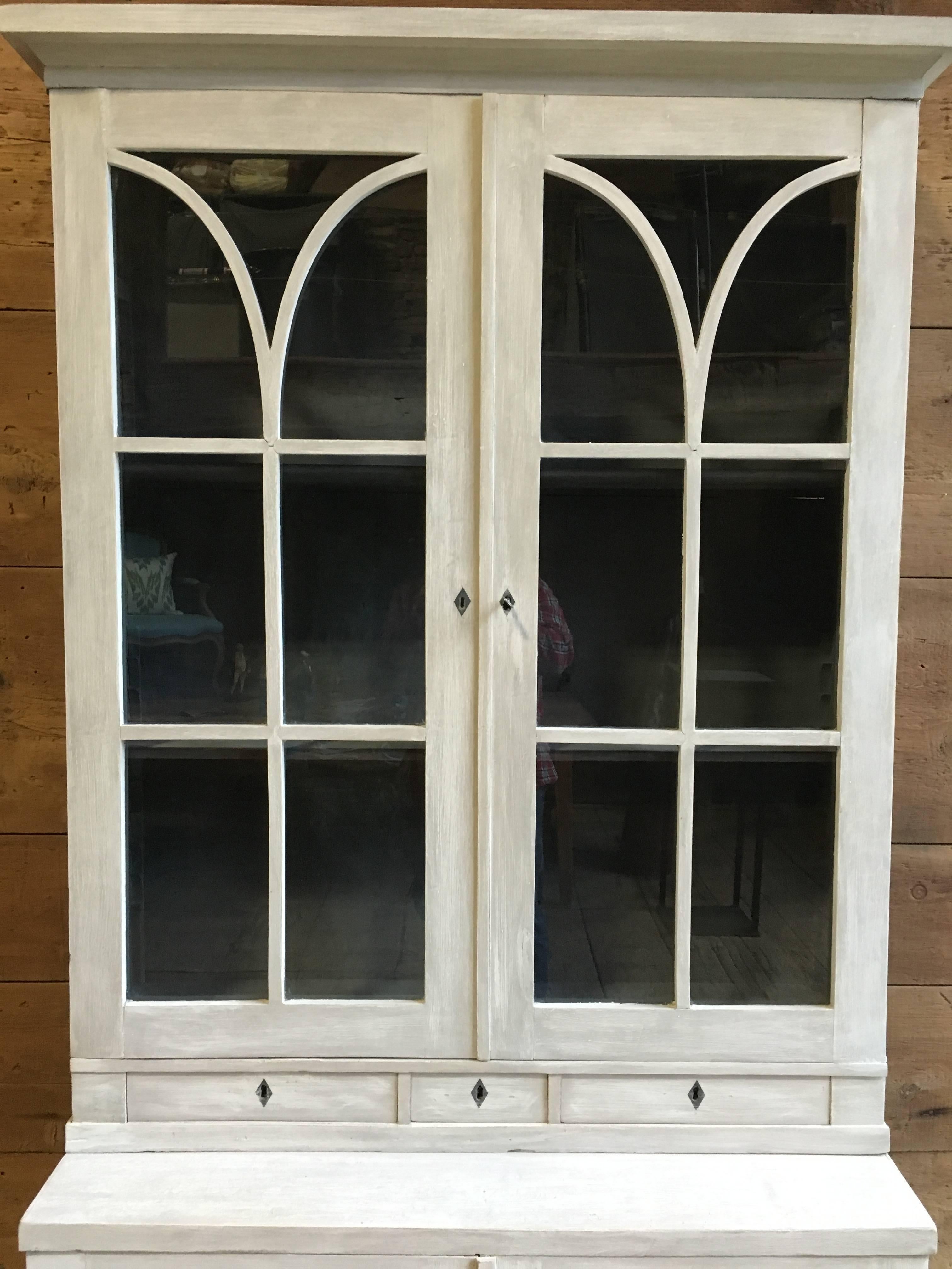 A French Directoire style grey painted cabinet or bookcase, in two parts, the upper part with glazed doors over three small drawers, with interior adjustable shelves and the lower cabinet with two paneled doors and interior shelves, early 19th