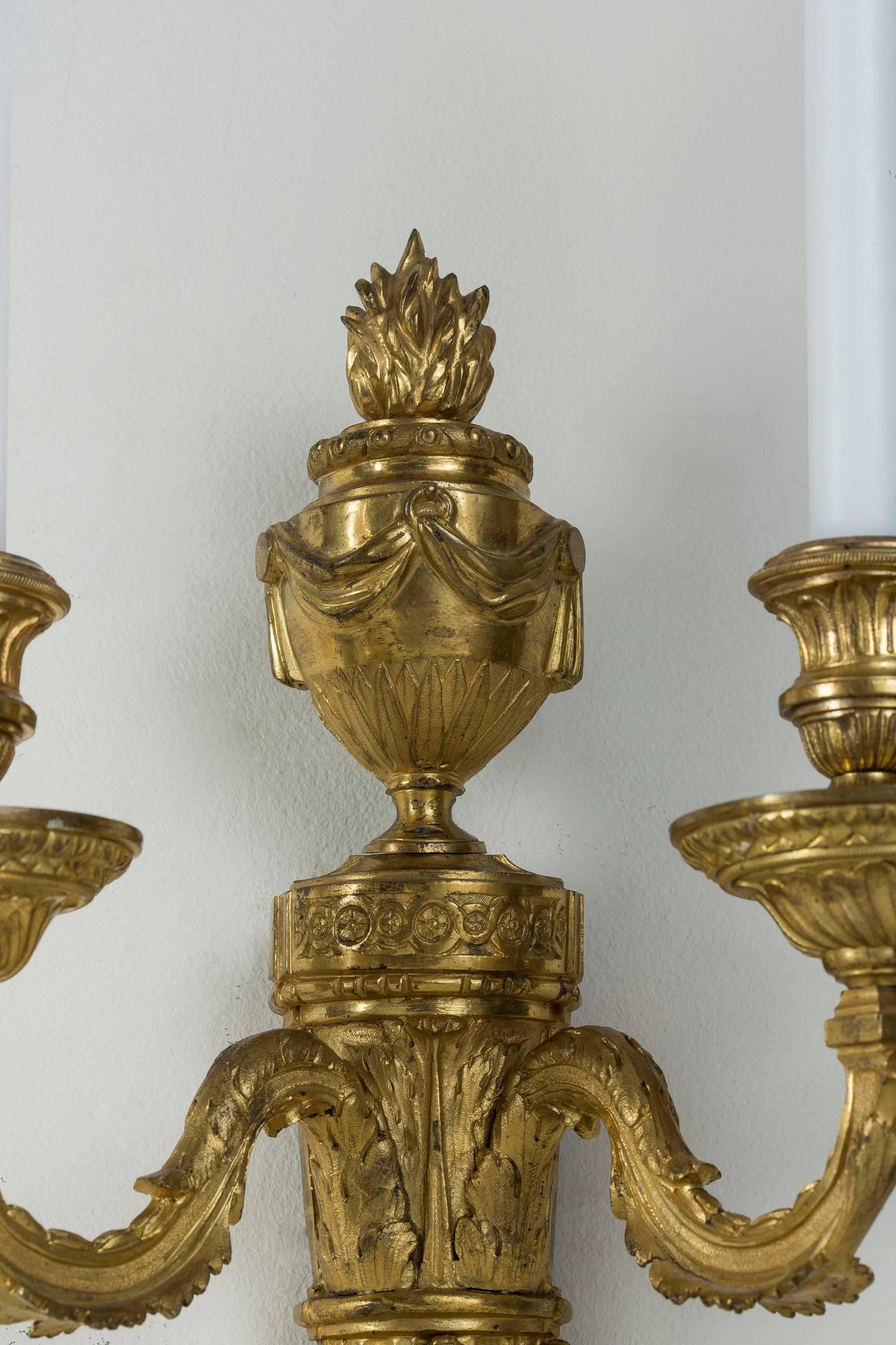 Each with a foliate-cast tapering fluted and backplate decorated with swags and surmounted by a swagged flaming classical urn, issuing foliate scrolled branches with gadrooned nozzle and foliate drip-pans. After a design by Jean-Louis Prieur.