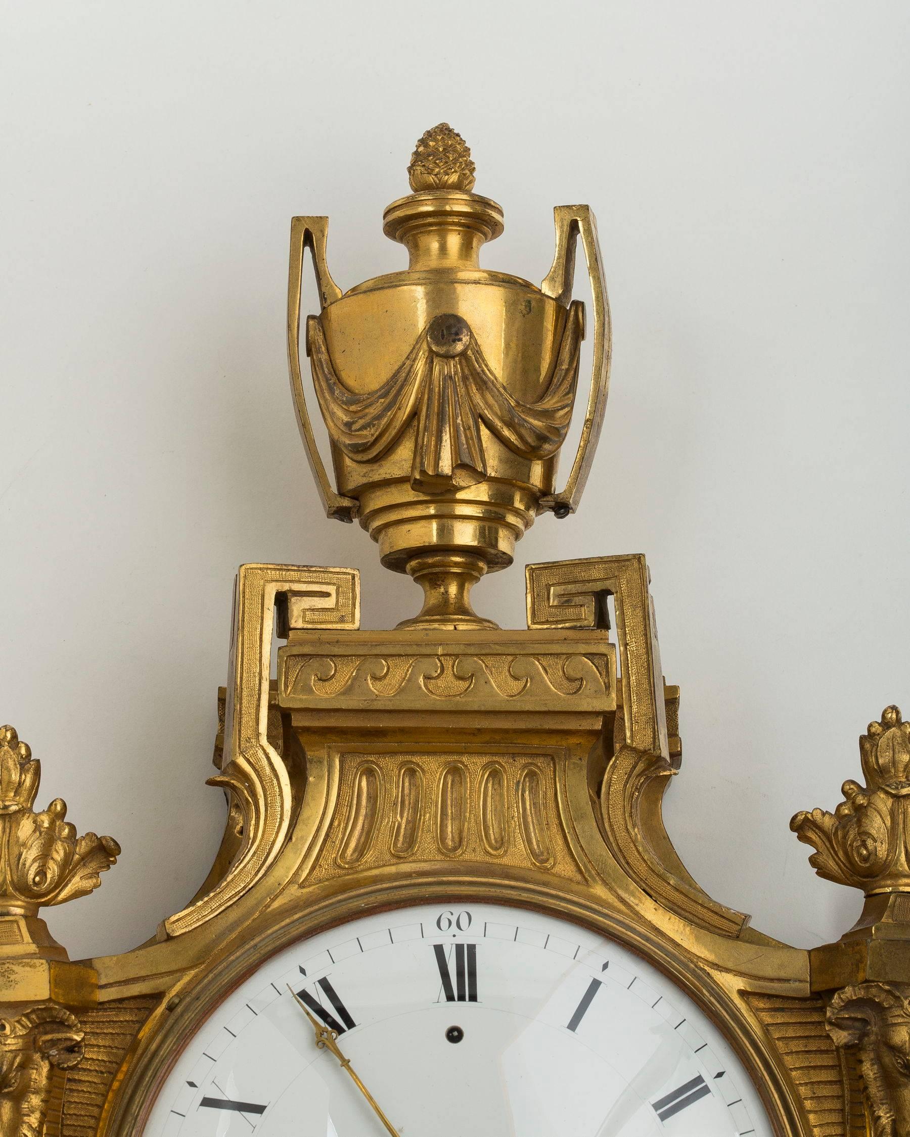 The circular glazed white enamel dial with Roman and Arabic chapters, set within a pierced case with ribbon-tied fruiting laurel trails, each side with a ram’s mask capped pilaster, surmounted by a twin-handled classical urn finial hung with swag,