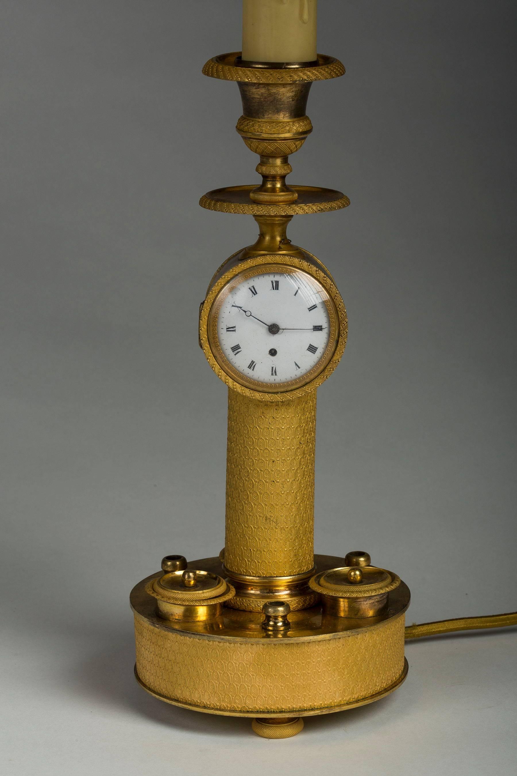 The circular base fitted with three inkwell compartments, the finely chased stem with an unmarked round clock and ending in a nozzle, mounted as a lamp.