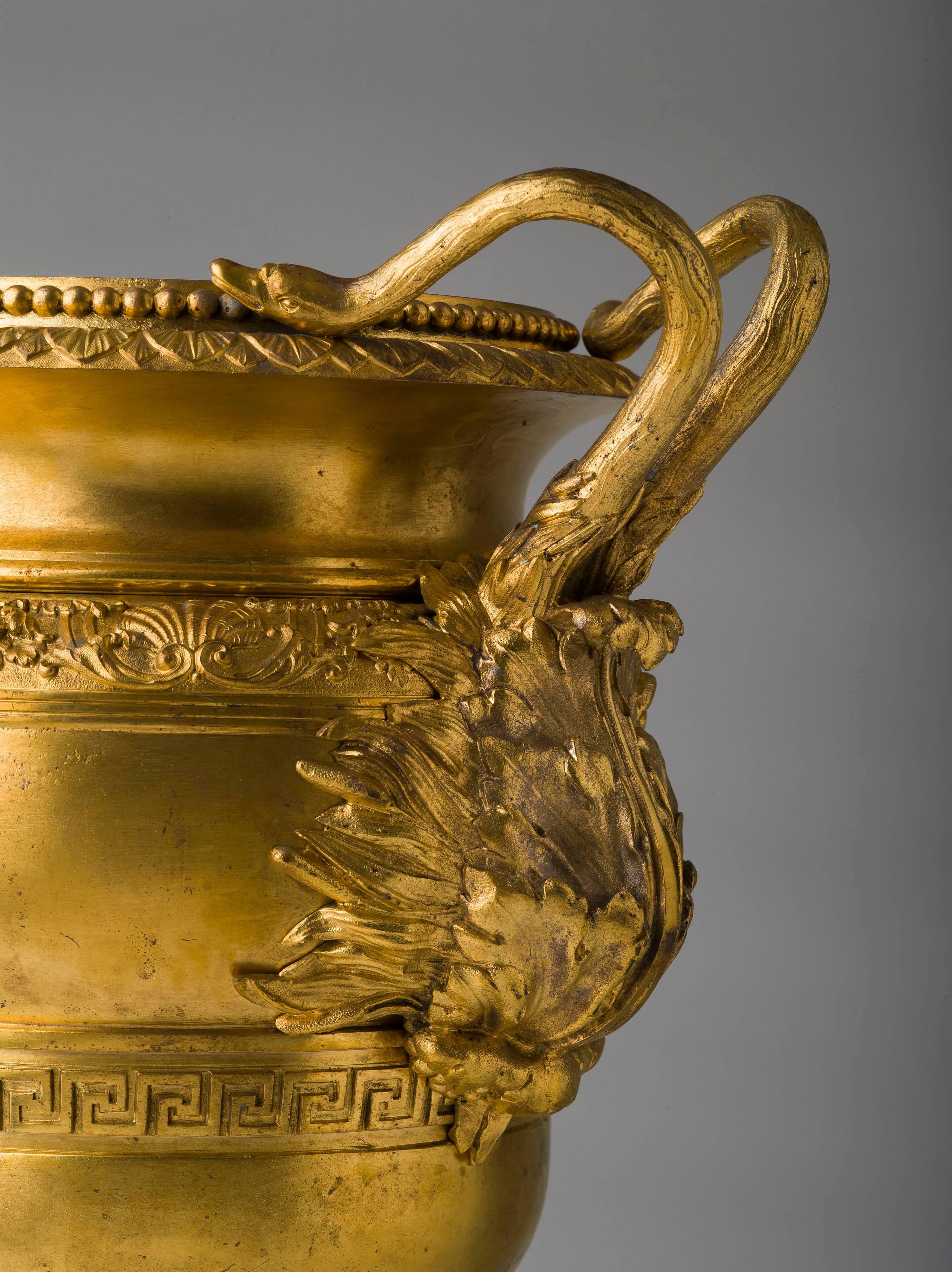 Of circular urn form with two double swan handles ending in acanthus, the body with two horizontal bands of shell and foliate and Greek key decoration, raised on a circular fluted foot ending in a square plinth. 

Following Matthew Boulton and