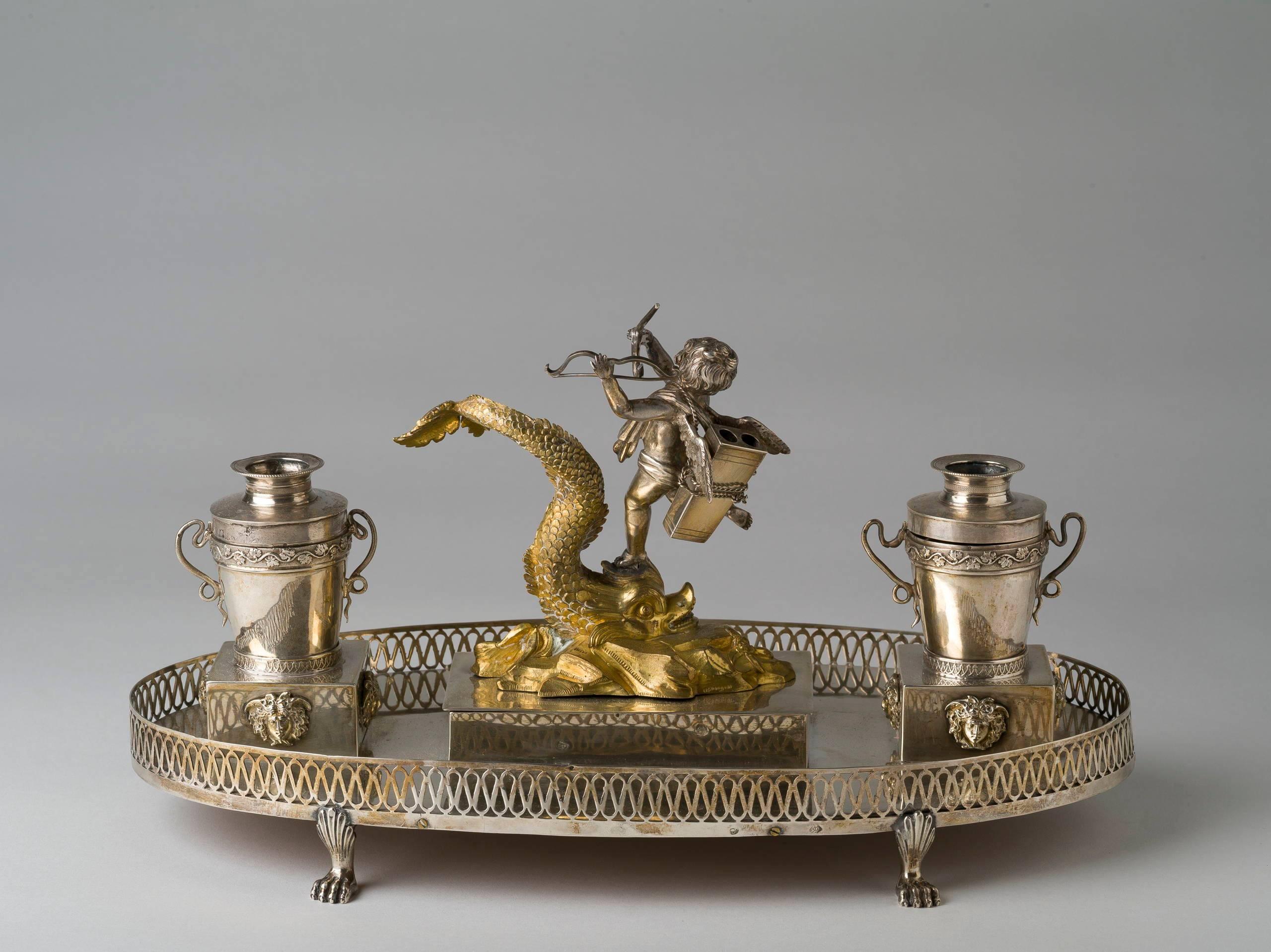 Of oval shape, with a cupid holding a bow and arrow riding aloft a gilt dolphin forming a lid over a compartment, flanked by ink and sand pots on masked plinths cast with Medusa masks and mounted to a lion's-paw footed oval galleried tray. Marked