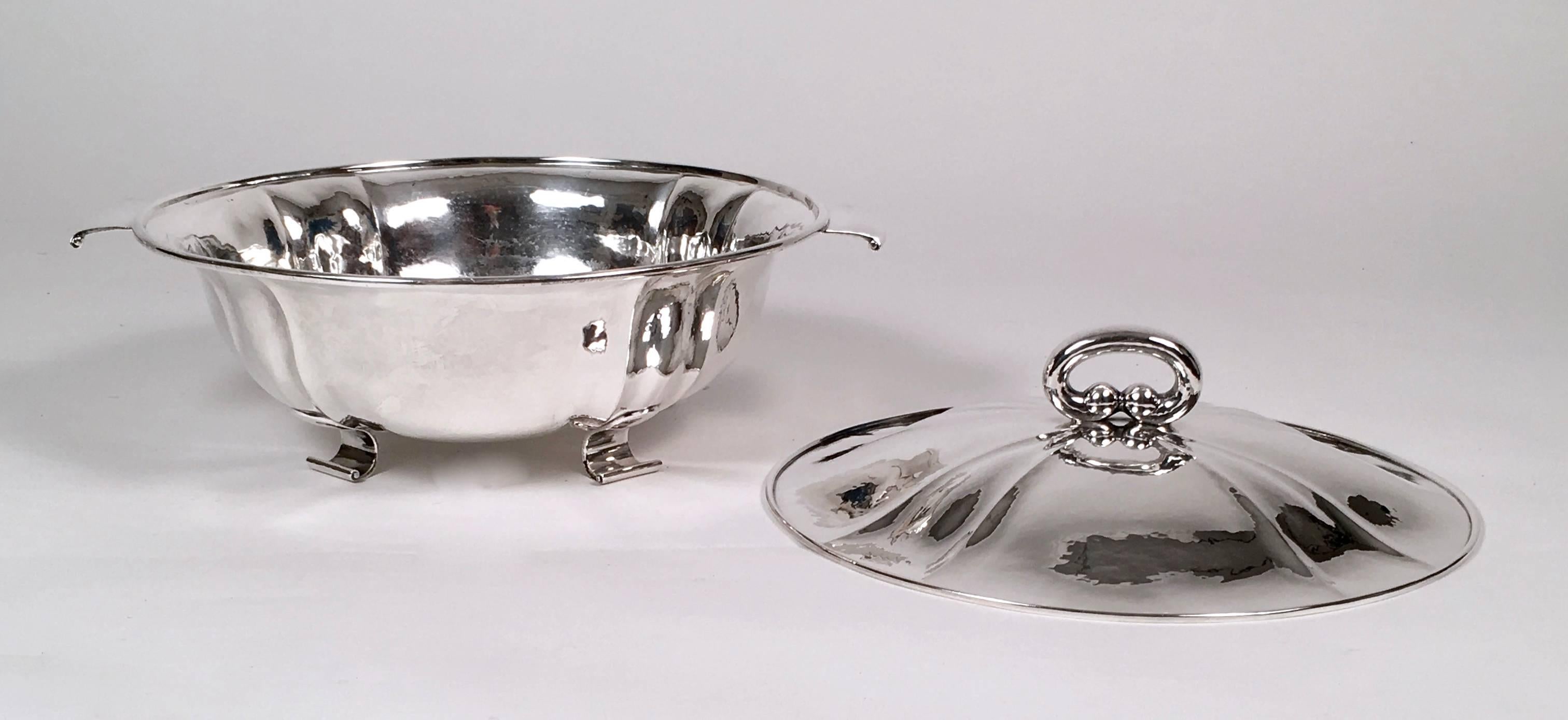 Sopera forrada de plata de ley suiza de gran calidad, hecha a mano, de forma circular, con tapa y base martilladas a mano, ambas con nervaduras decorativas, asa de lazo ovalada en la tapa y asas y patas de correa aplanadas con roleos. Firmado