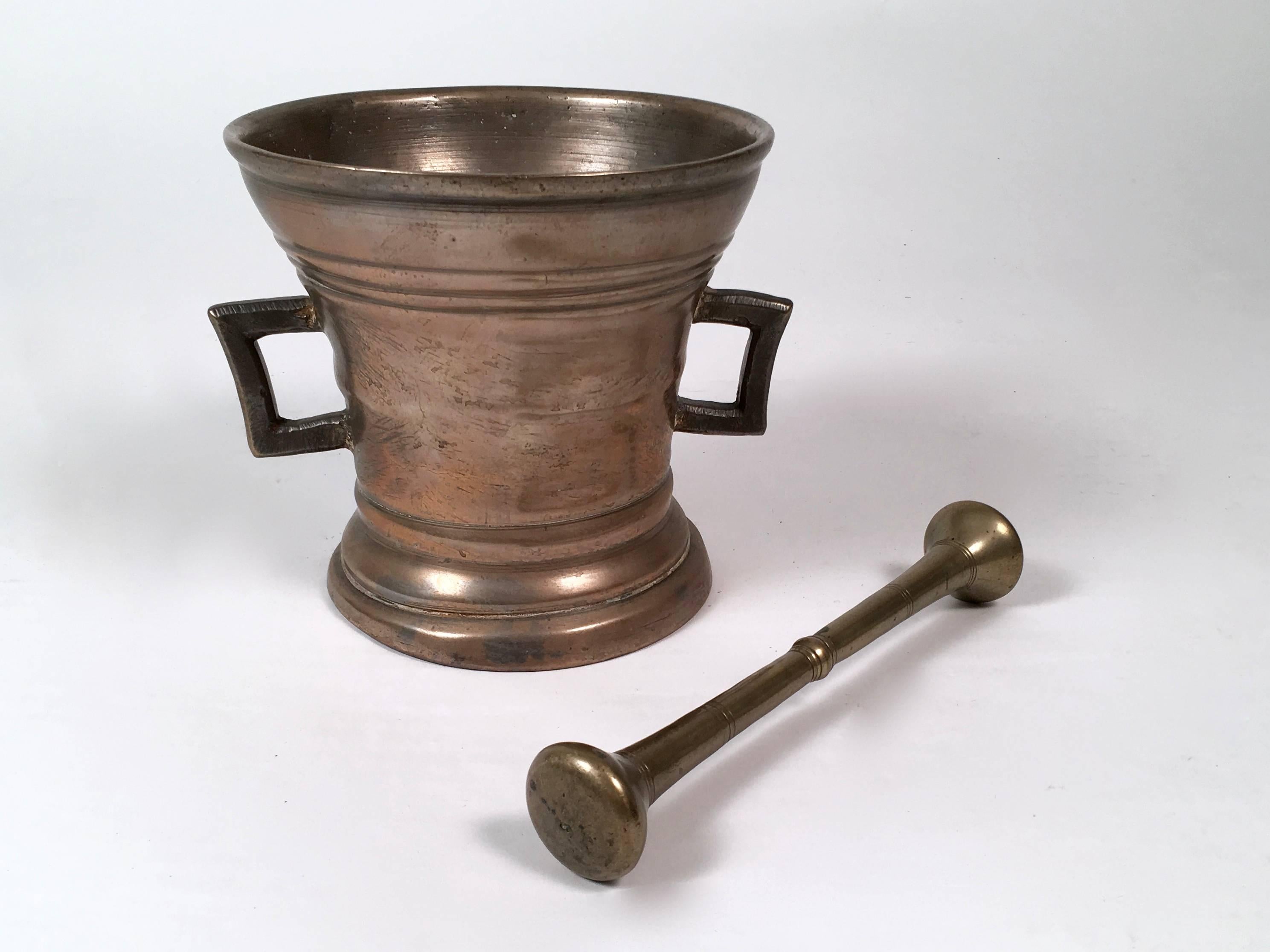 An early European cast bronze apothecary mortar and pestle of graduated cylindrical form, with ribbed decoration, on a circular spreading base, with rectangular strap handles. Great weight and quality. Found in Switzerland.

Measures: Overall