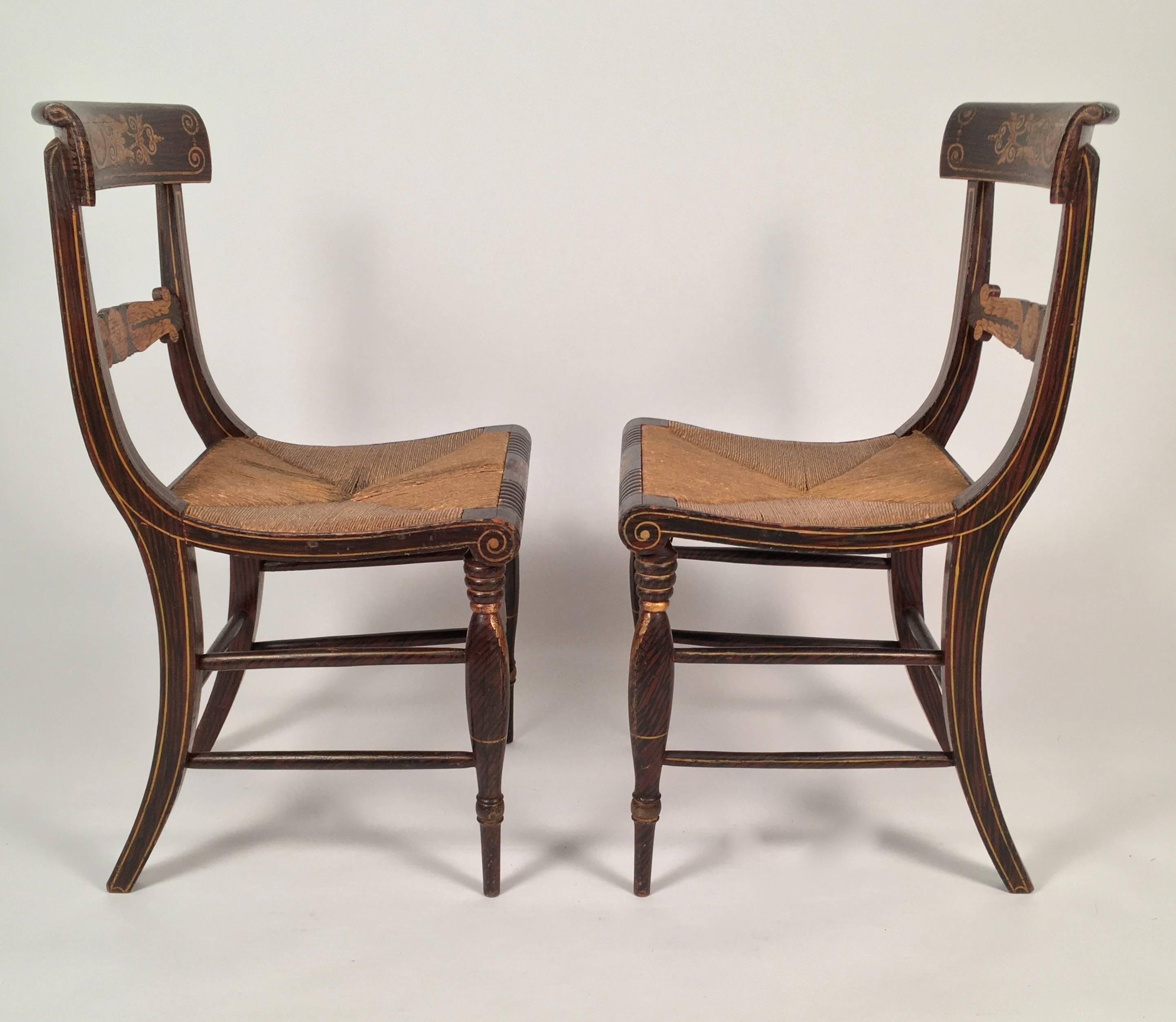 A pair of Federal period neoclassical fancy painted Klismos chairs, retaining their original stenciled gilded decoration, depicting rosettes, anthemion, acanthus leaves, scrolled and pinstripe decoration over grain painted rosewood, with rush seats