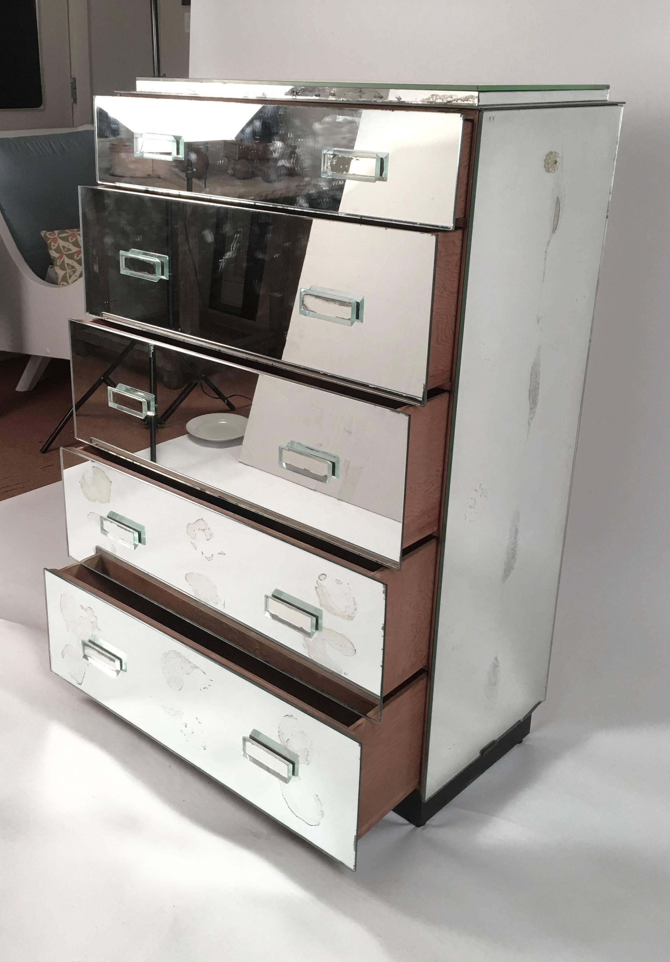 An Art Deco mirrored glass dresser of tall rectangular form with five graduated drawers, each with two mirrored glass rectangular pulls on a recessed black painted base. Generous storage, and versatile for many types of interiors.