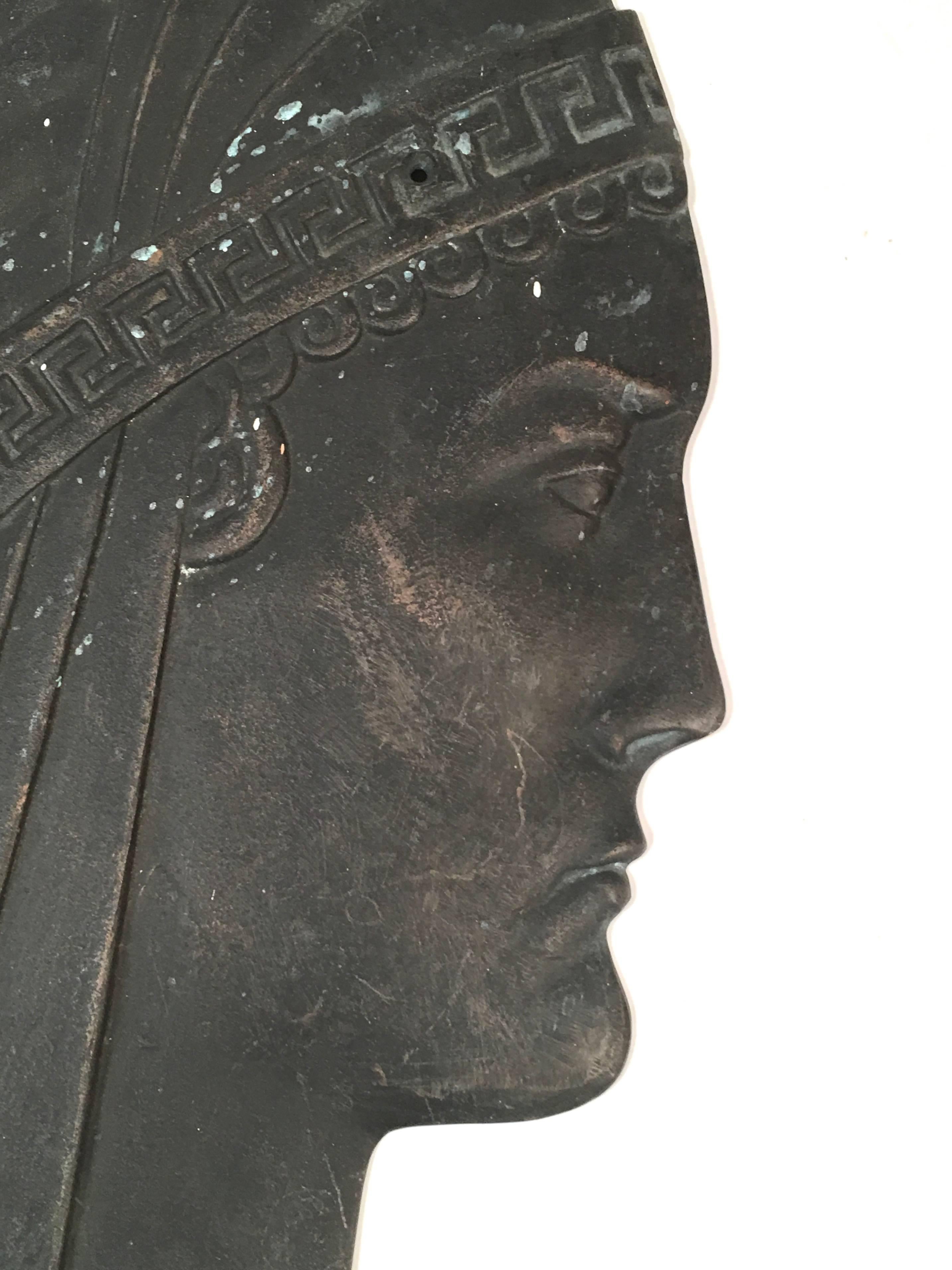 An Art Deco period cast iron neoclassical style head of a Grecian woman, in bas relief, likely an architectural ornament or wall decoration from a Hollywood Regency interior in black cast iron, the woman's head with stylized features, a Greek key