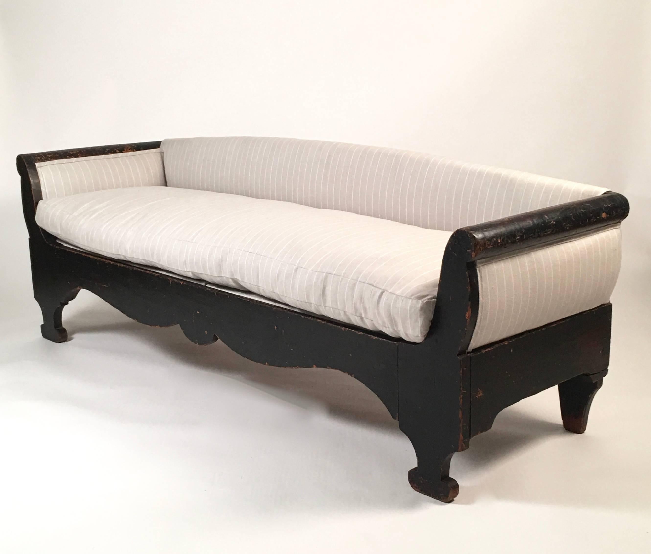 An American country sofa, or upholstered bench, of unusual, wonderfully sculptural form, in black painted wood with scrolled arms and scalloped front rail, raised on compressed oval feet. This sofa has been newly reupholstered in a high quality