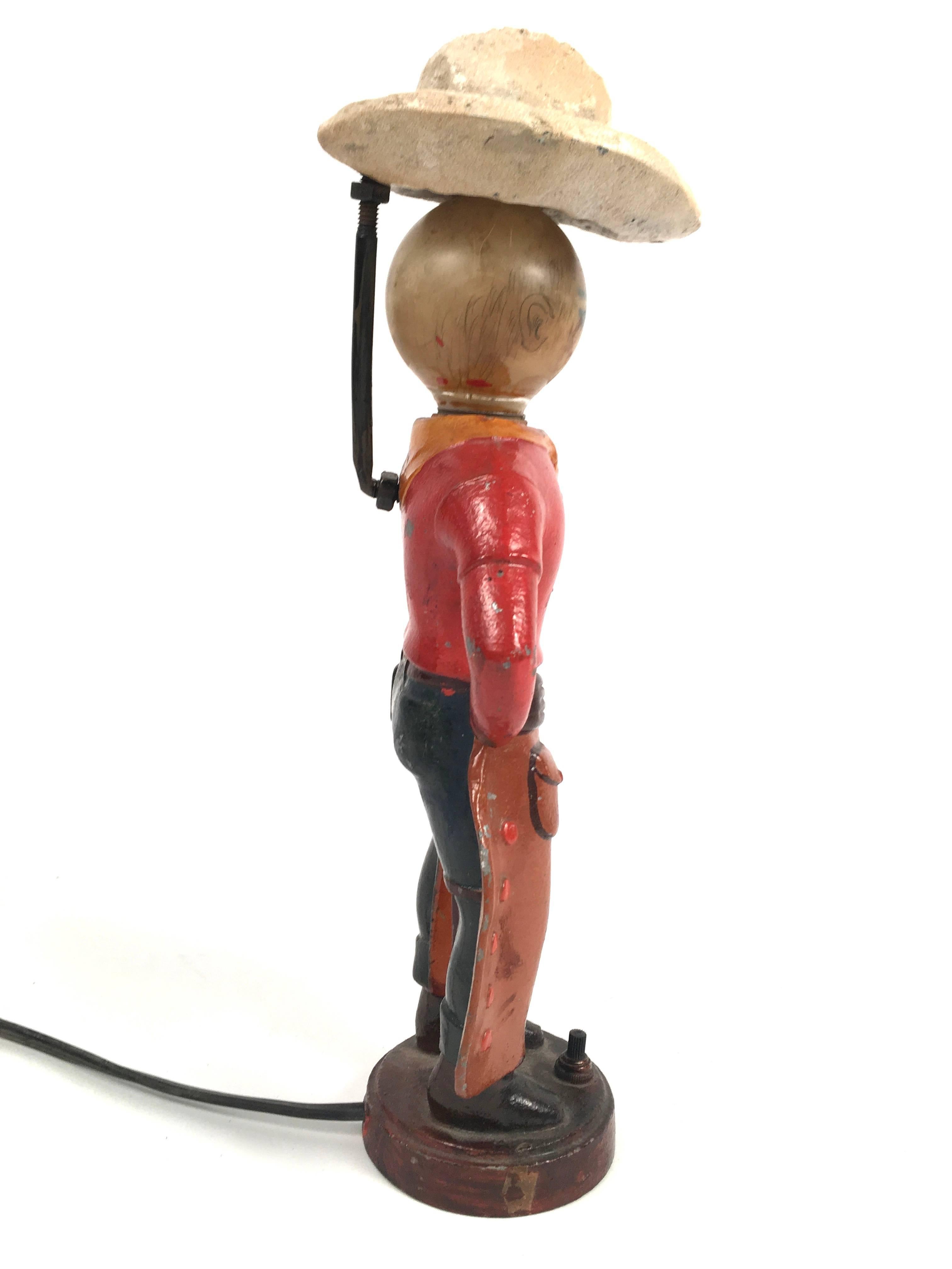 A playful painted cast iron vintage figural cowboy lamp, American, circa 1930s, with a hat that pivots sideways for accessing the lightbulb which is hand decorated with facial features, the cowboy with hands on his hips, wearing a red shirt with