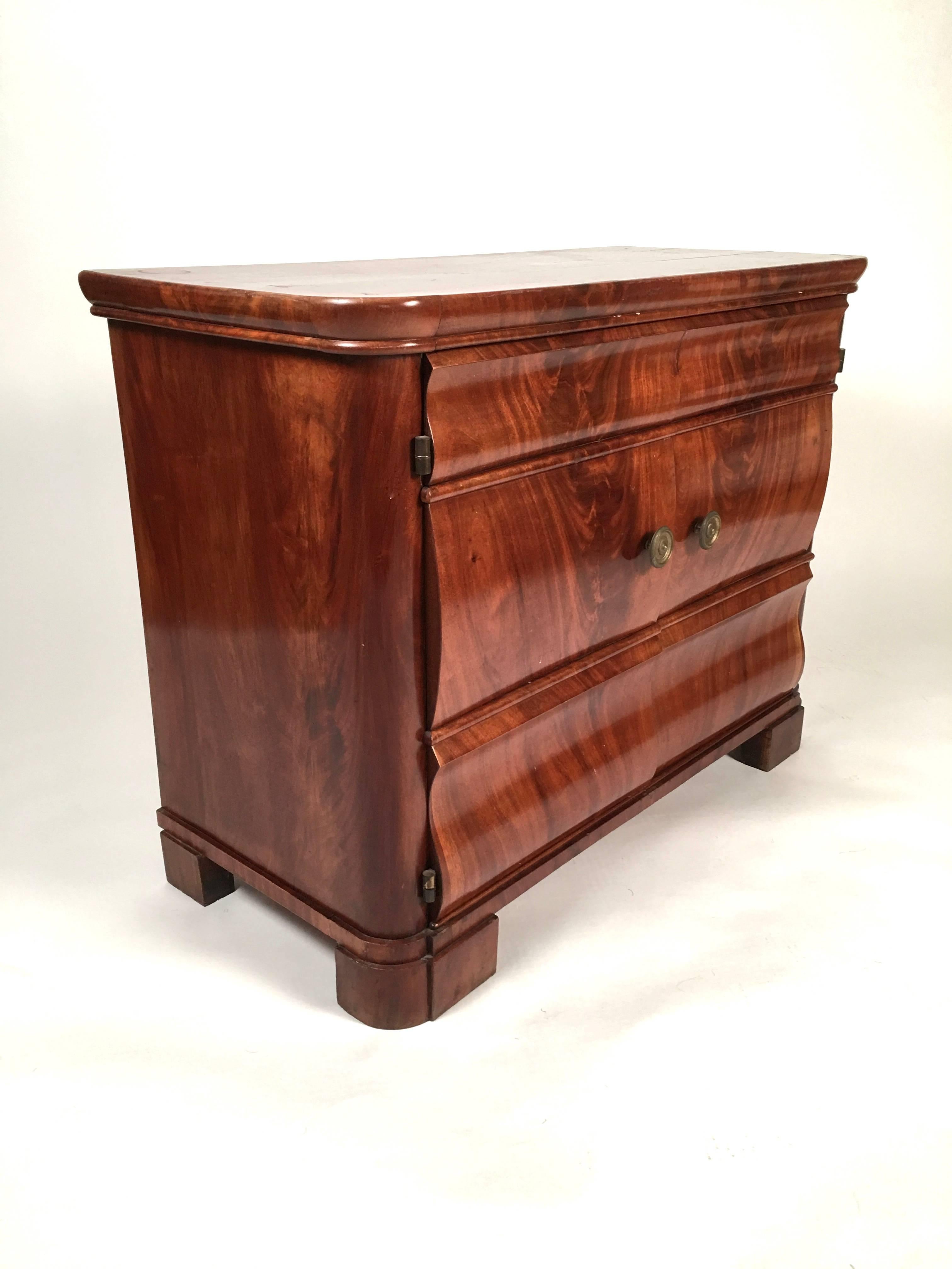A Biedermeier mahogany two door side cabinet, of wonderful small scale, the serpentine carved doors in richly figured mahogany, with 2 brass neoclassical round door pulls, the rectangular top with curved corners, all raised on bracket feet.