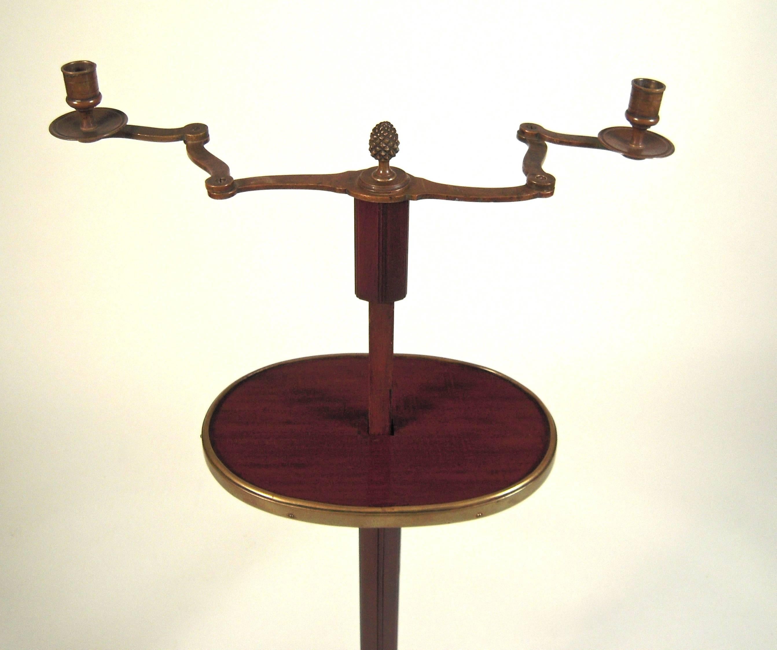 An unusual, charming and quirky Edwardian, adjustable height, two-light mahogany and brass candle Stand or drinks table, English, circa 1910, with brass pineapple finial over two adjustable candle arms above a brass bound mahogany oval shelf,