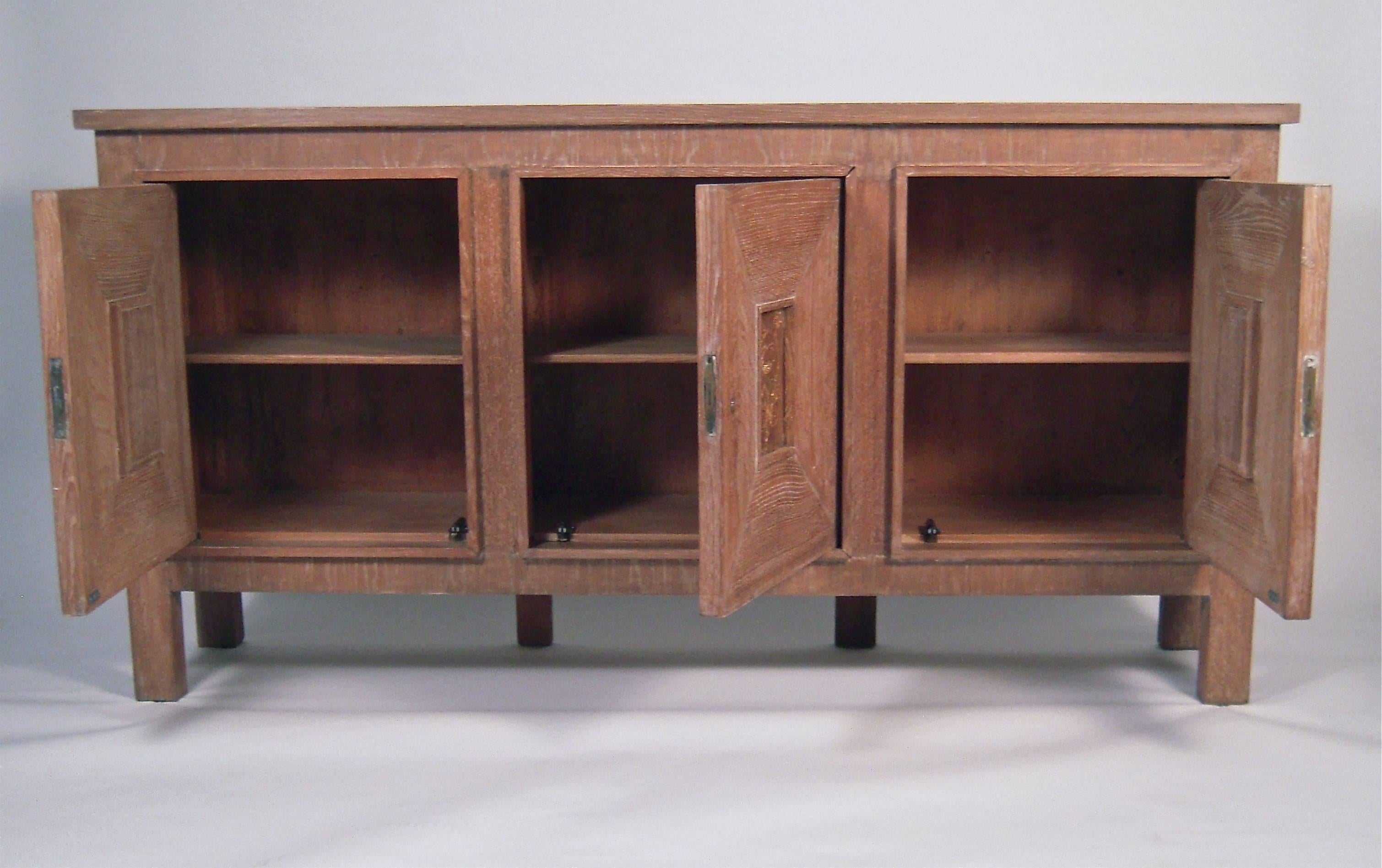 A well proportioned cerused, oak credenza, French or American, circa 1940s, of rectangular form, the paneled cupboard doors each centered by an incised, gilded decorative floral and figurative panel, raised on square section legs. Doors are fitted