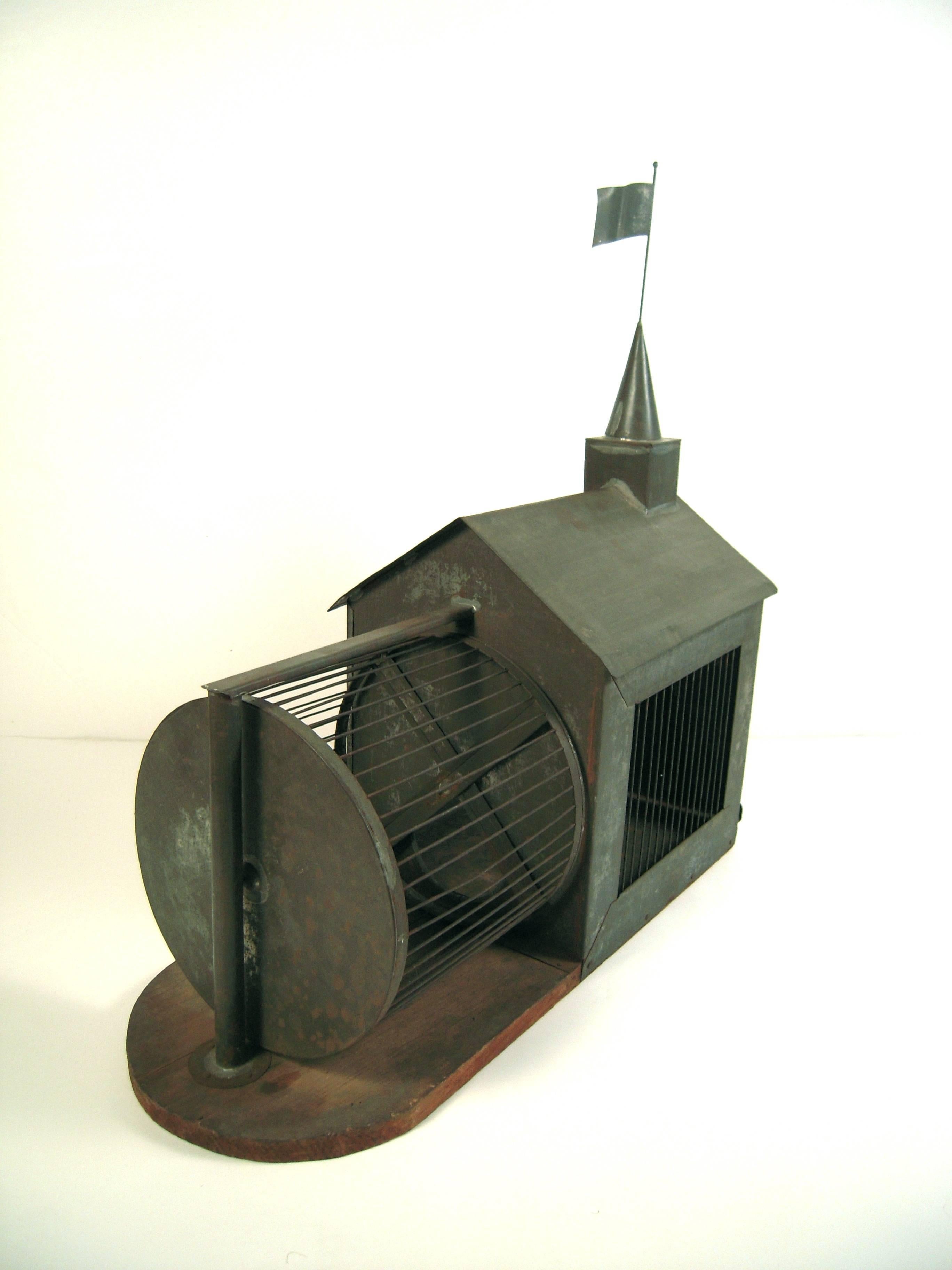 A rare, complete 19th century American Folk Art architectural squirrel cage, beautifully preserved, in tin in the form of a meeting house with conical tower and flag waving in the wind, mounted on a wooden base. The squirrel cage has a sliding door,