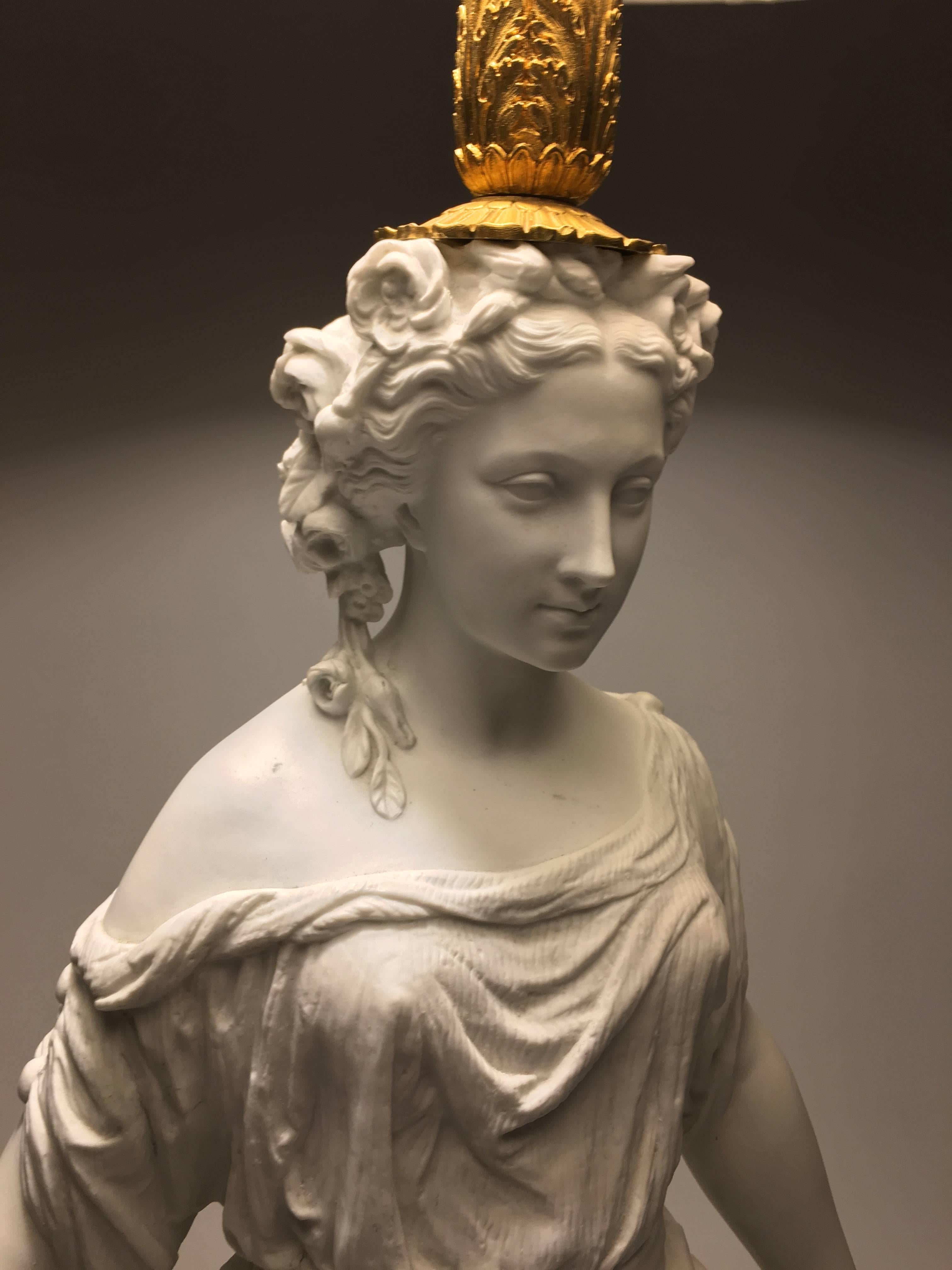 A fine quality French neoclassical lamp in biscuit porcelain in the form of a classical female figure, wearing a beautifully modelled draped garment, holding a scroll in each hand, on a fine gilded base with beaded and berried laurel wreath