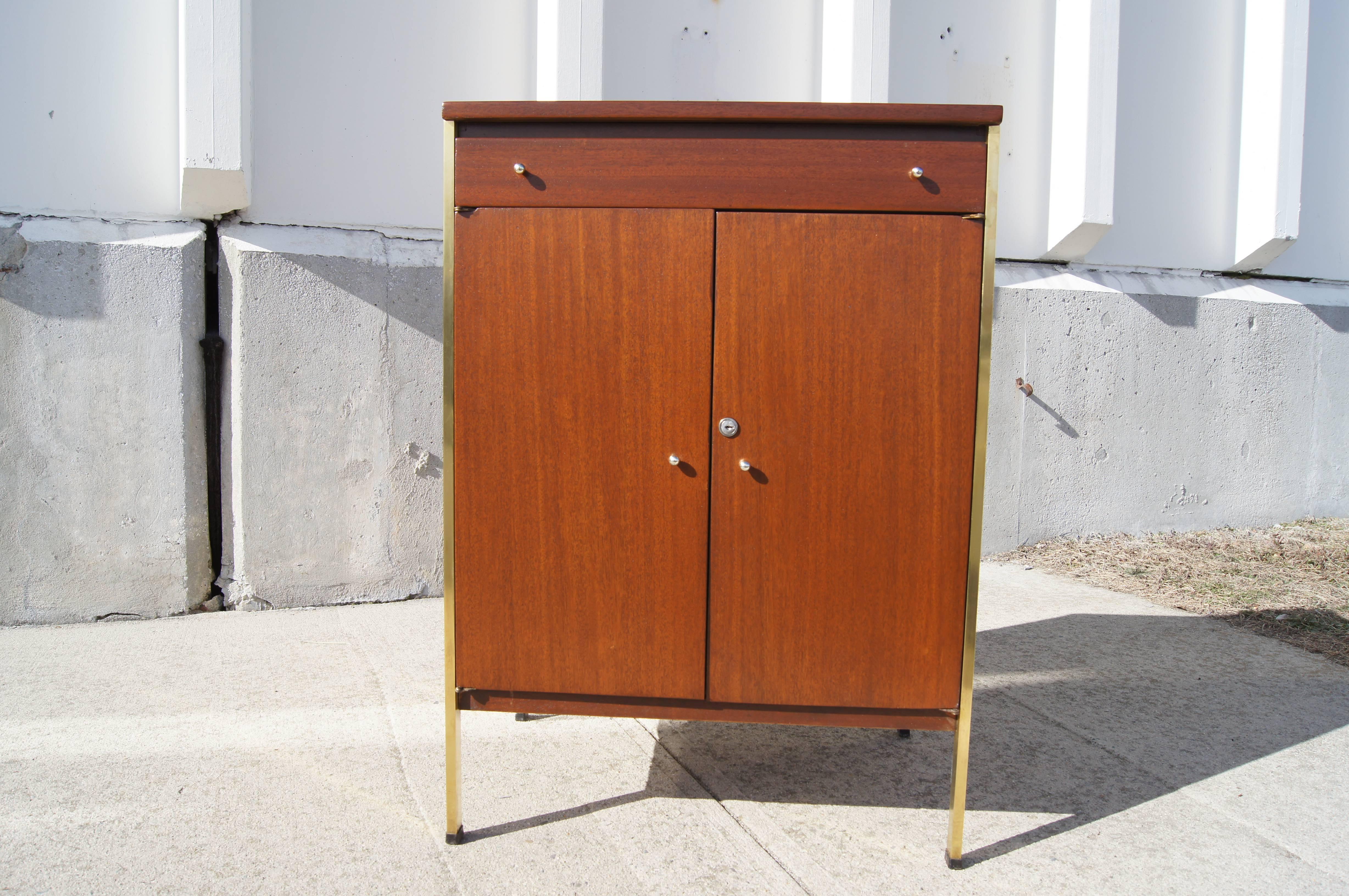 Paul McCobb designed this small mid-century cabinet for the Calvin Furniture Company. The walnut-stained mahogany case stands on brass rails that extend up the sides. Below a shallow drawer two doors conceal storage space with an adjustable shelf,