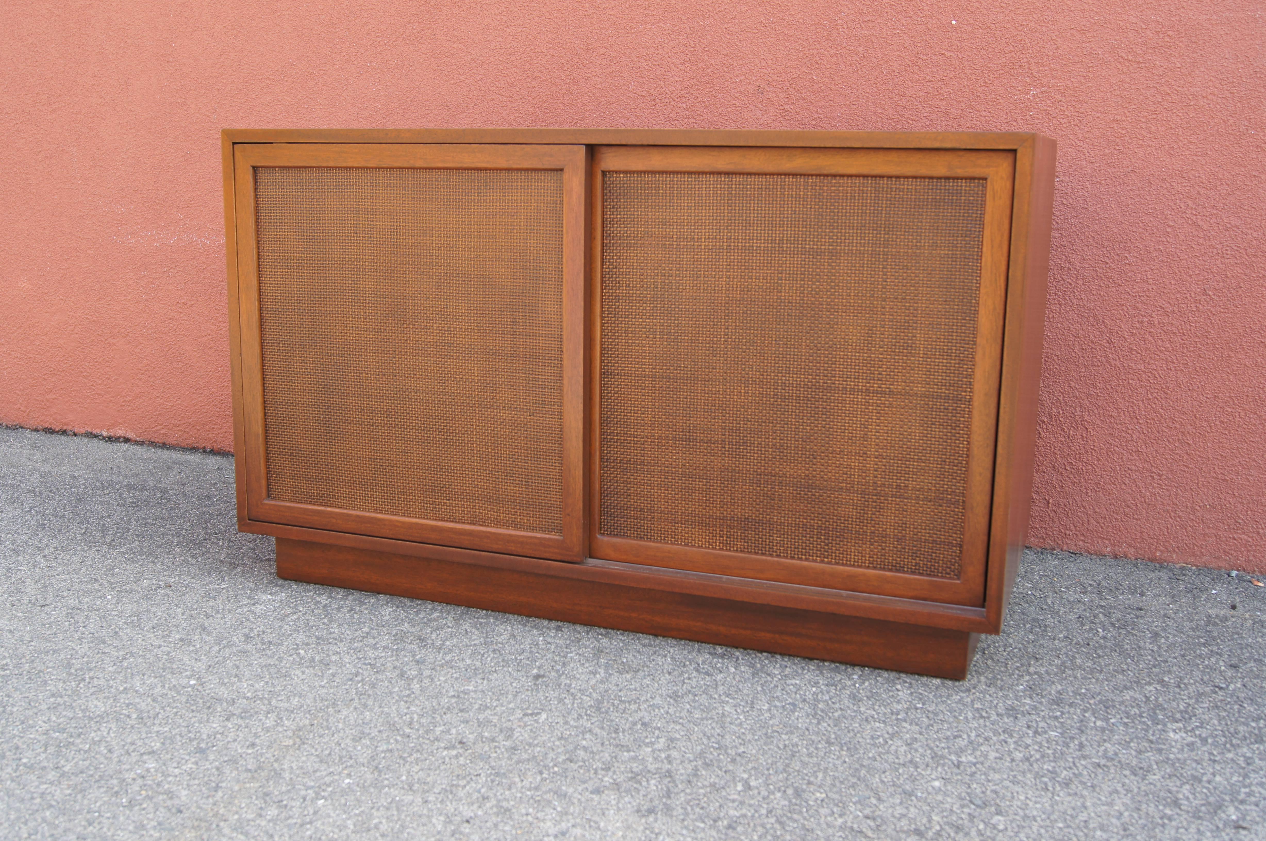 Designed by Harvey Probber, this small midcentury cabinet features a mahogany case on a plinth base. Cane-front sliding doors conceal, on the left, three deep drawers with white lacquered fronts and, on the right, additional adjustable