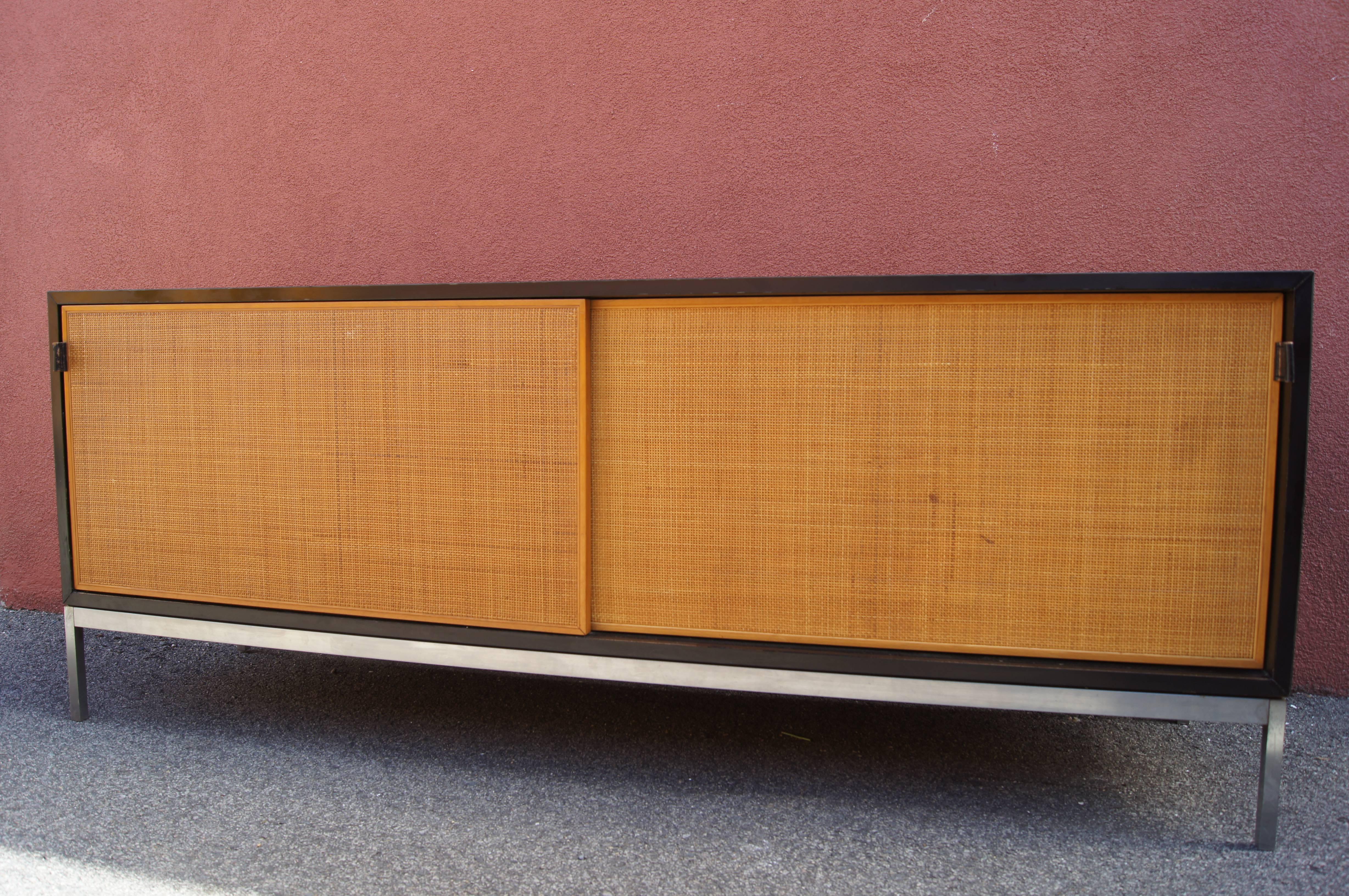 Dieses Sideboard ist ein Beispiel für die klassische, geradlinige Ästhetik von Florence Knoll. Das Gehäuse aus schwarzem Laminat steht auf einem Stahlsockel und verfügt über hübsche Schiebetüren aus Schilfrohr mit Ledergriffen. Im Inneren befinden